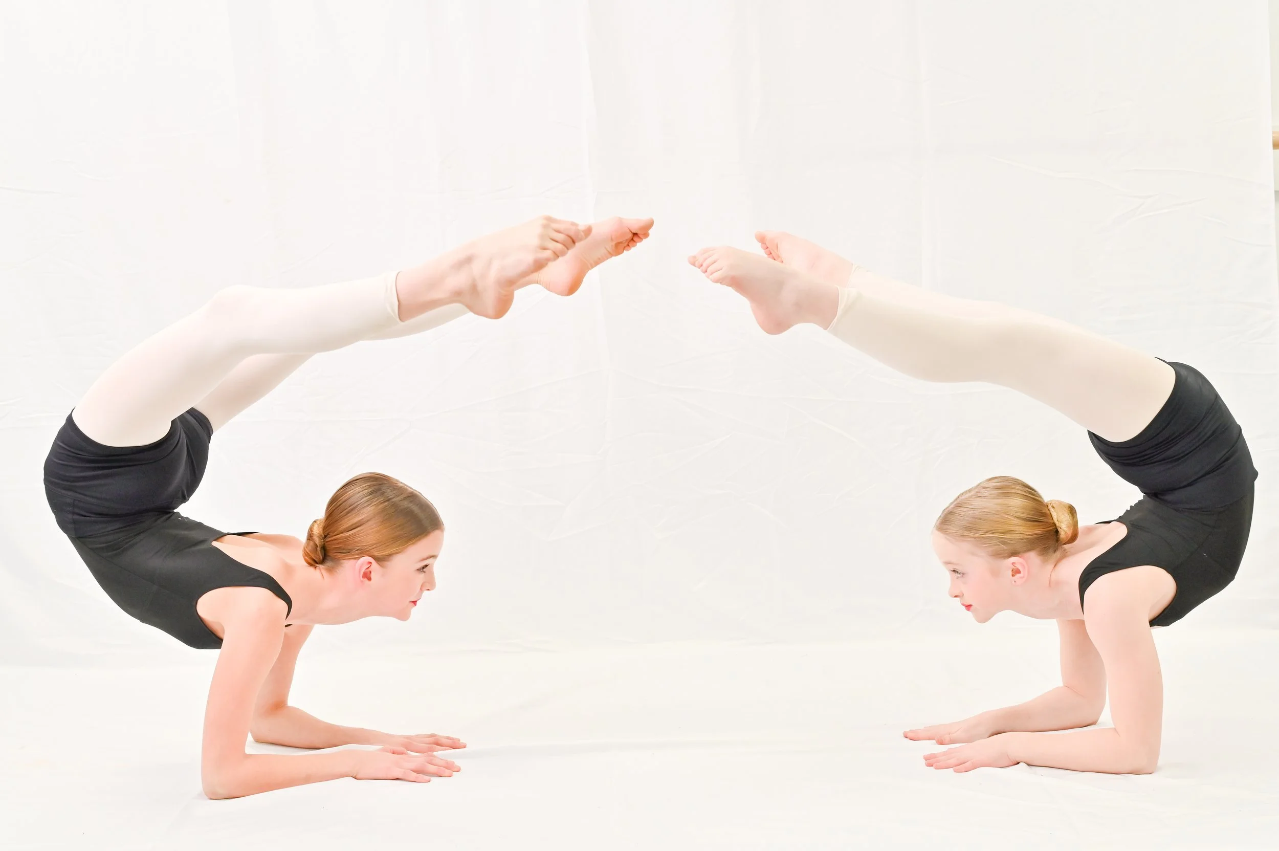 Two women with red hair and black leotards practicing yoga in a headstand pose with their fingers interlocked, facing each other against a white background.