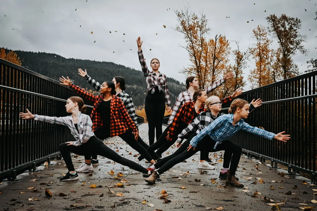 A group of young women wearing plaid shirts performing yoga stretches on a bridge with fall foliage and mountains in the background.