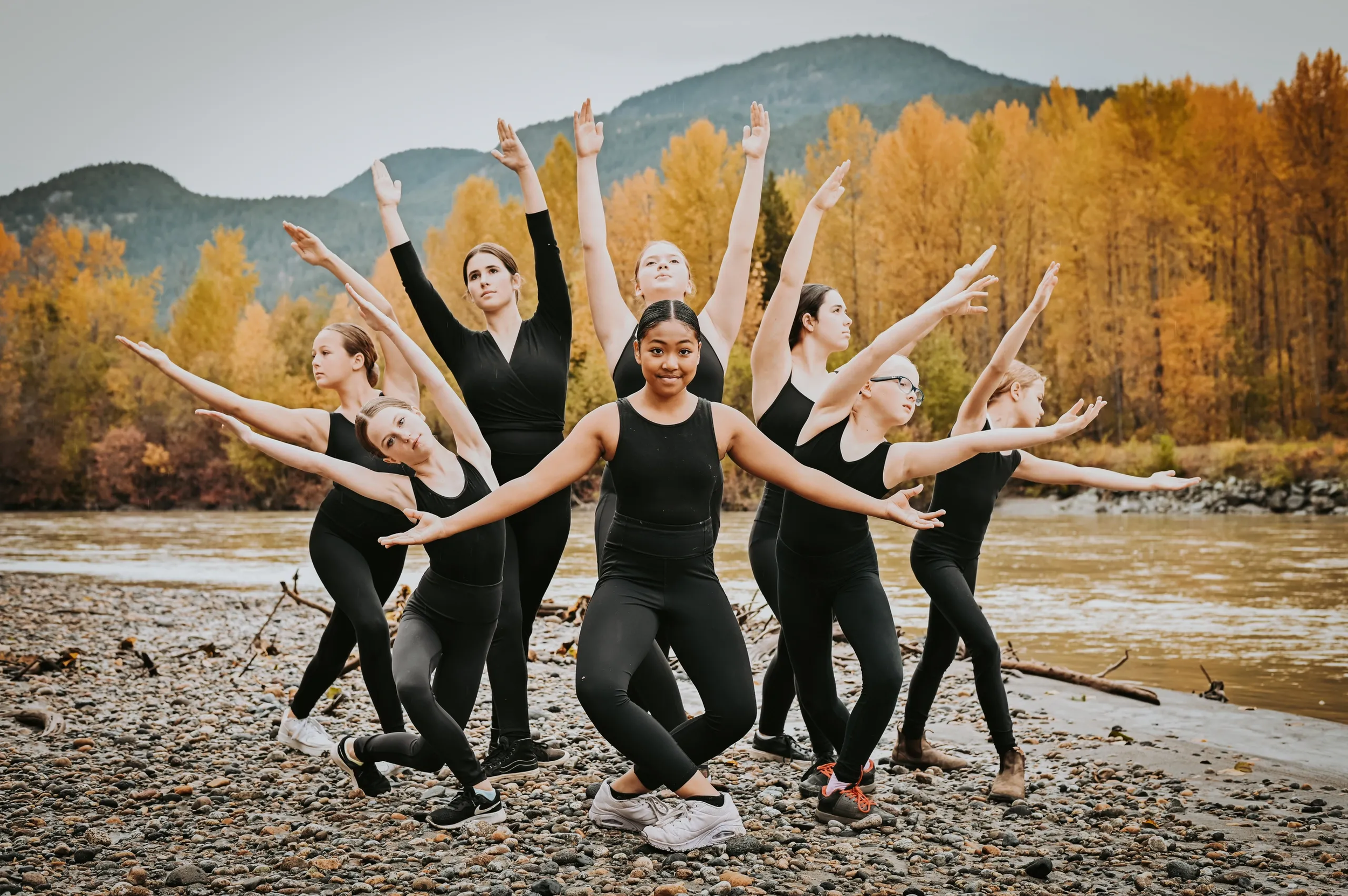 Group of young women practicing ballet outdoors on rocky beach during autumn, with colorful trees and distant mountains in background.
