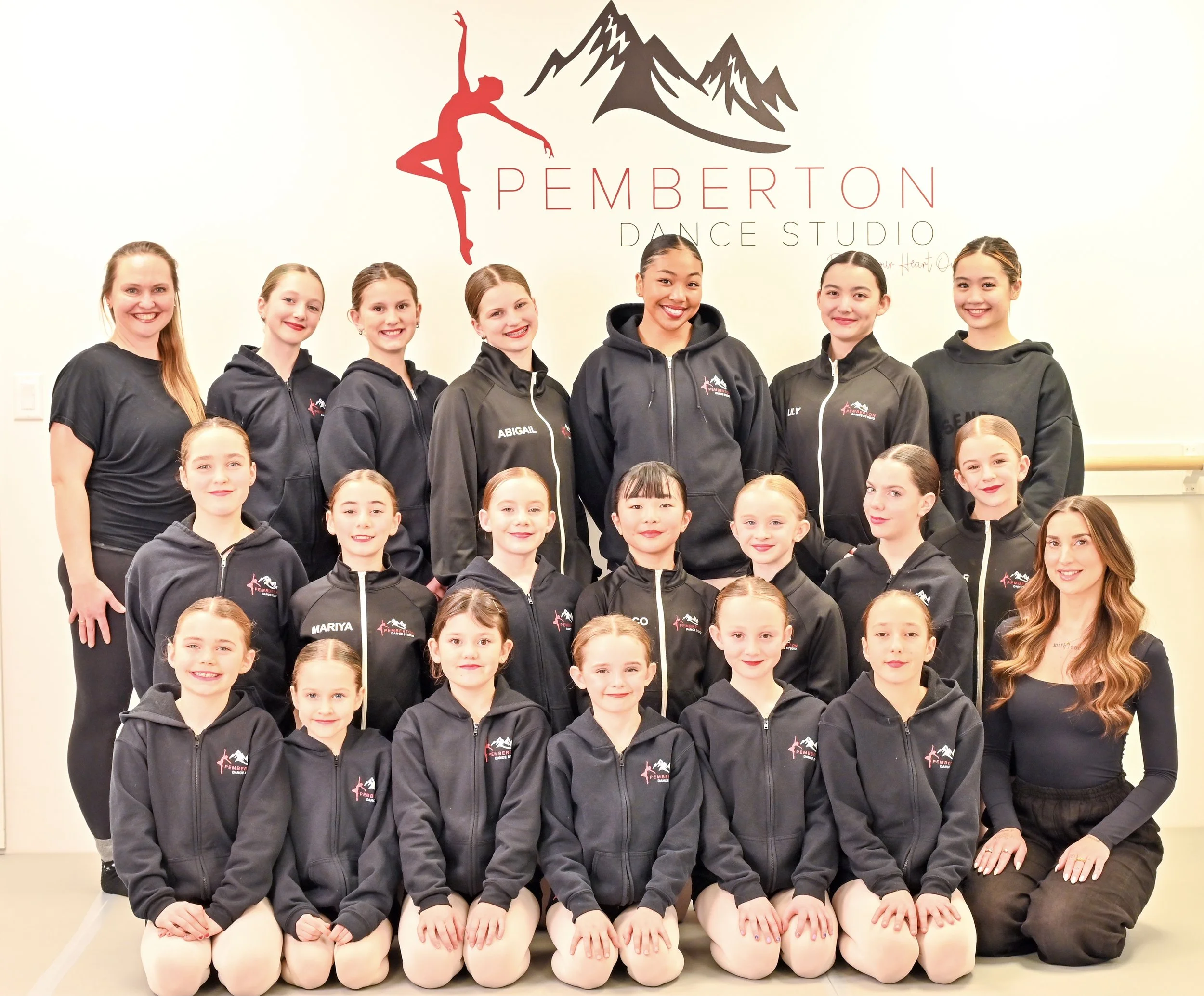 Group of young ballet dancers and their instructor in a dance studio, wearing black hoodies with a mountain and dancer logo, smiling for a group photo in front of a wall with a dance studio logo.
