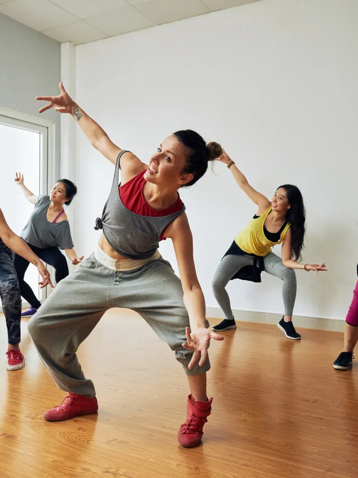 Group of women participating in a dance or fitness class indoors, with one woman in the forefront striking a dynamic pose.