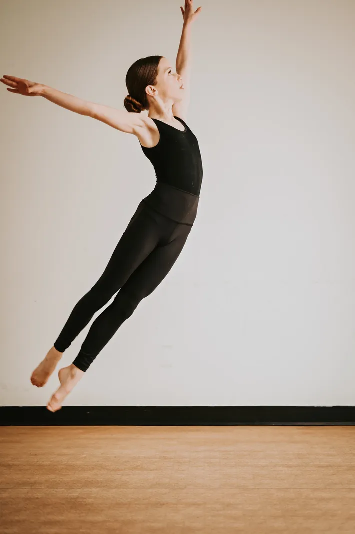A woman in black workout clothes appears to be levitating or jumping against a plain wall.