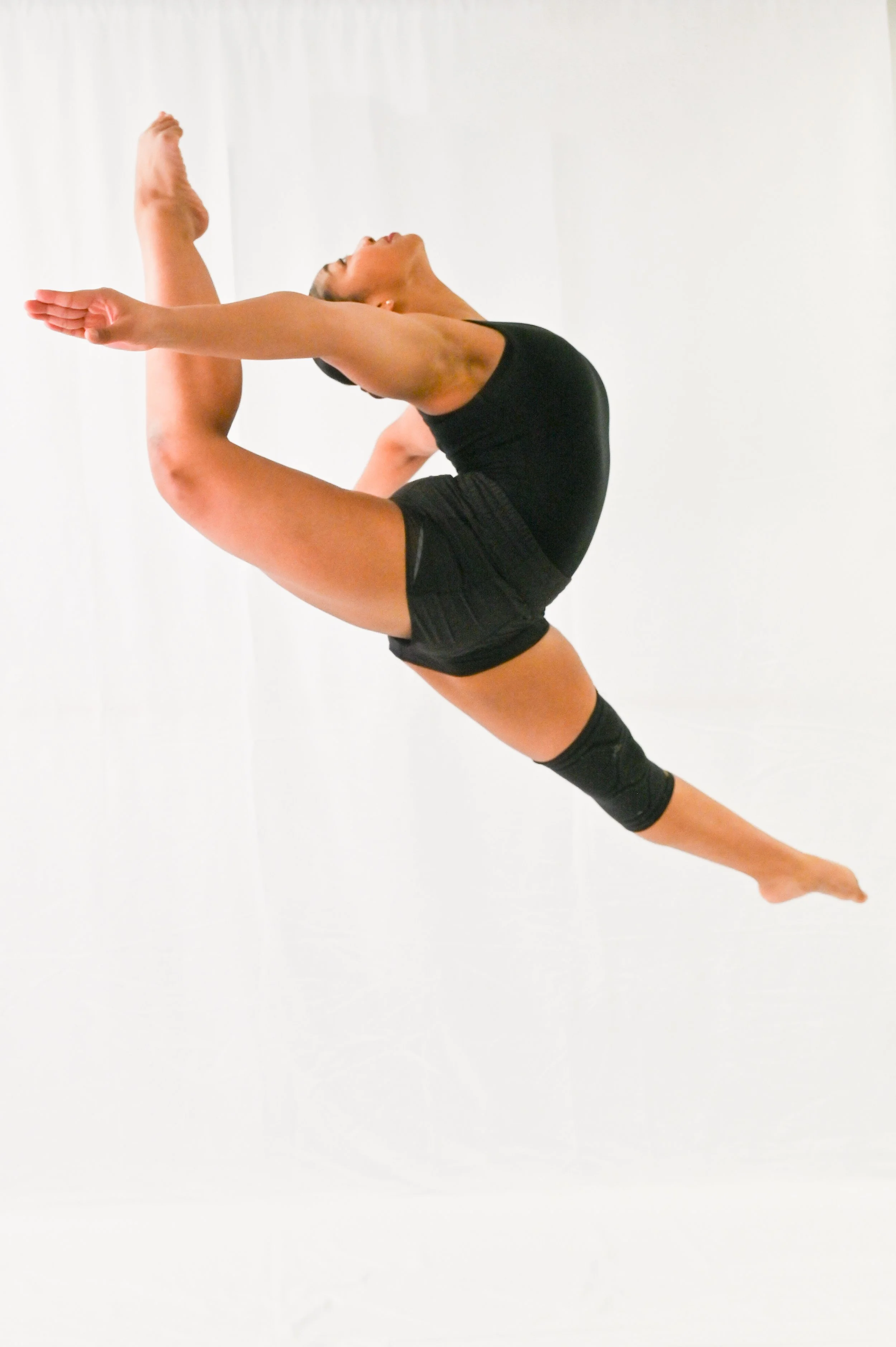 A female gymnast in black workout attire performing a split leap with arms extended forward and legs split in the air against a plain white background.