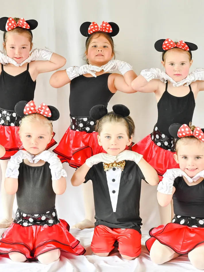 Six young children dressed in Minnie Mouse costumes, with red skirts, black tops, white gloves, and Minnie Mouse ears with red polka dot bows, posing together.