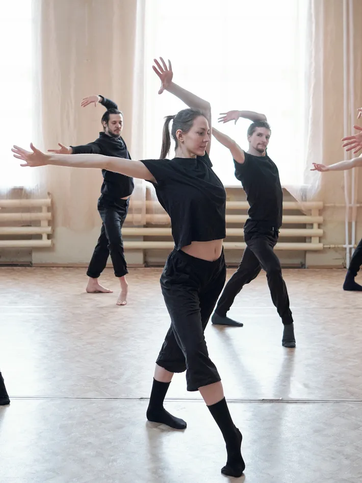 Group of dancers practicing ballet or contemporary dance in a dance studio with large windows and wooden floor.
