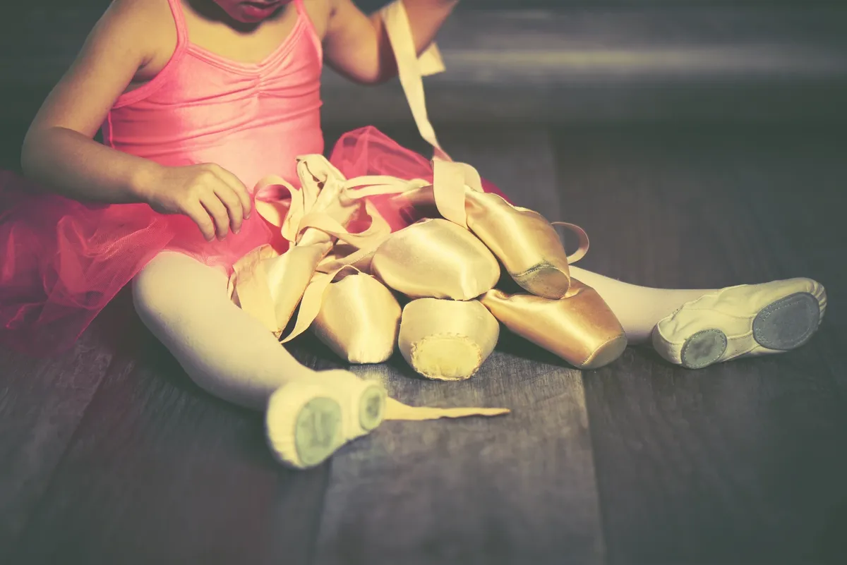 A young girl wearing a pink ballet tutu and ballet slippers sits on a dark floor, gathering gold ballet slippers in her lap.