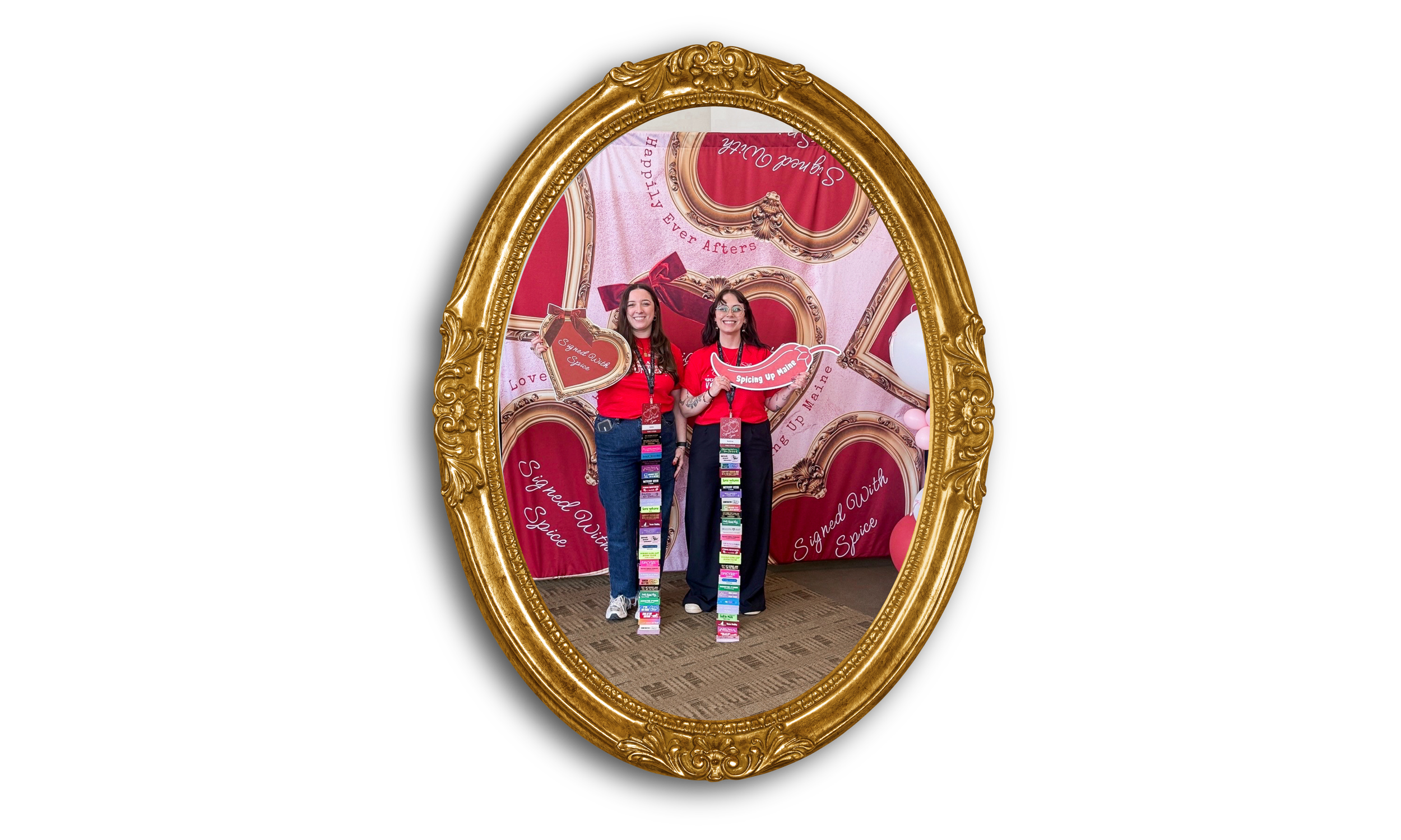 Two women smiling inside a gold ornate frame, holding themed signs, at a display with a pink and red backdrop with decorative frames. They wear red shirts with event lanyards.
