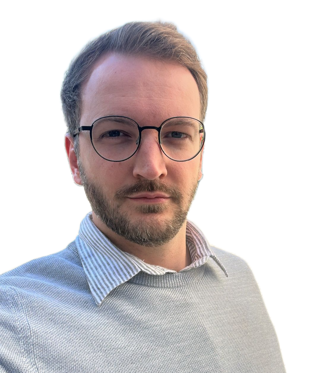 A young man with light brown hair, glasses, and a beard, wearing a collared shirt and a sweater, taking a selfie outdoors.