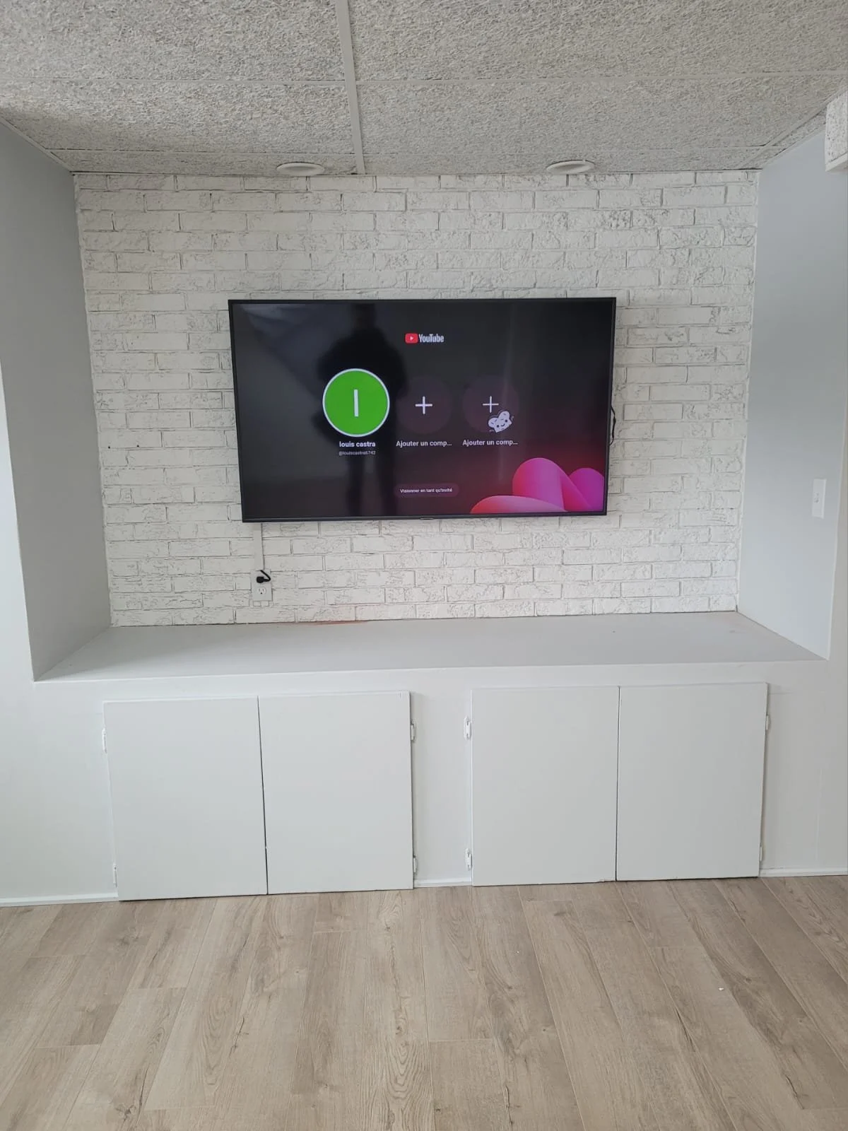 Un écran de télévision monté sur un mur en briques blanches, dans un salon moderne avec un mobilier blanc en bas, et un sol en bois clair.