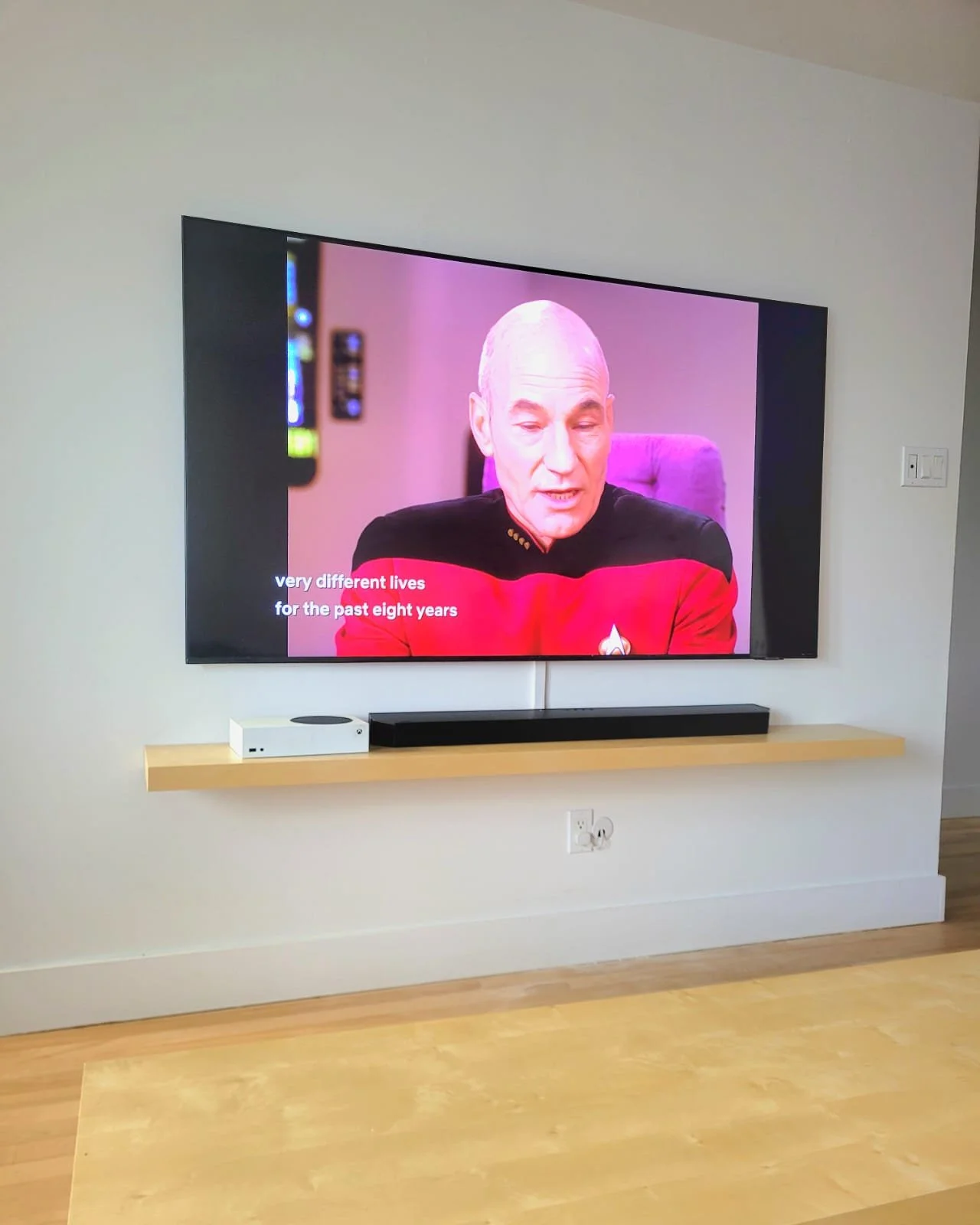 Une télévision affichant une scène d'une personne dans un uniforme de Star Trek, avec des sous-titres en anglais qui disent "very different lives for the past eight years". La télévision est placée sur un mur clair avec une étagère en dessous, suppor