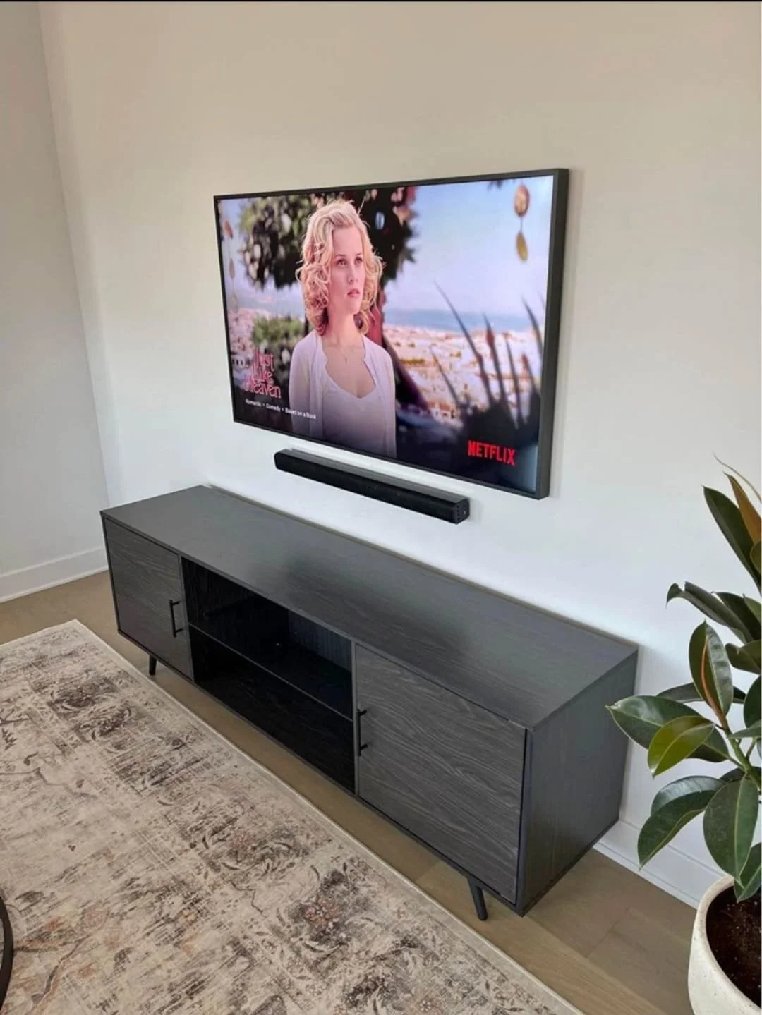 Écran de télévision fixée au mur affichant une scène d'une femme blonde dans un paysage en plein air, avec un bar de son surround en dessous. Meuble en bois gris sous la télévision, plante verte à droite, tapis beige et motifs sombres sur le sol.