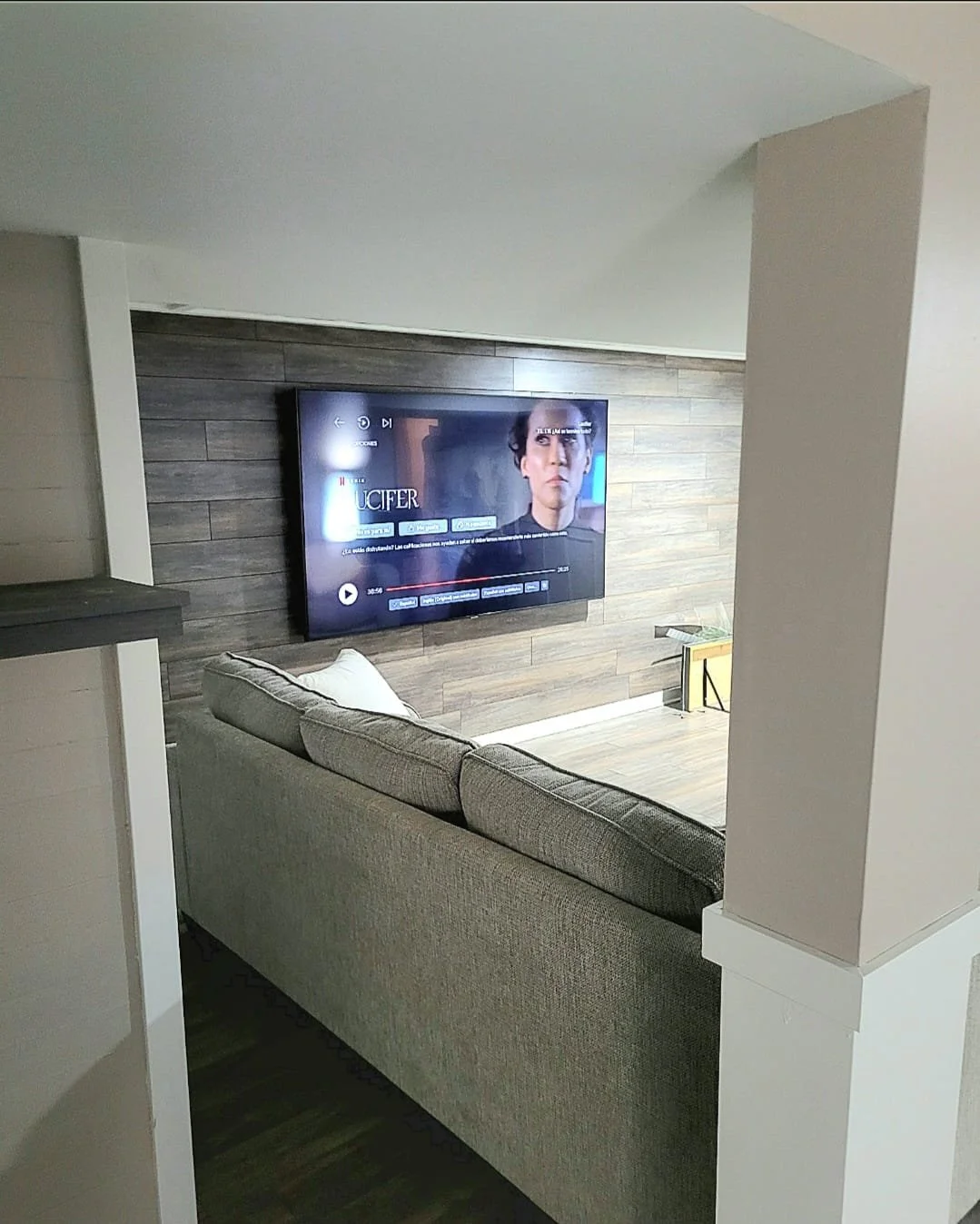 Salon avec un mur en bois, une télévision murale et un canapé gris, vue partielle à travers un espace ouvert.