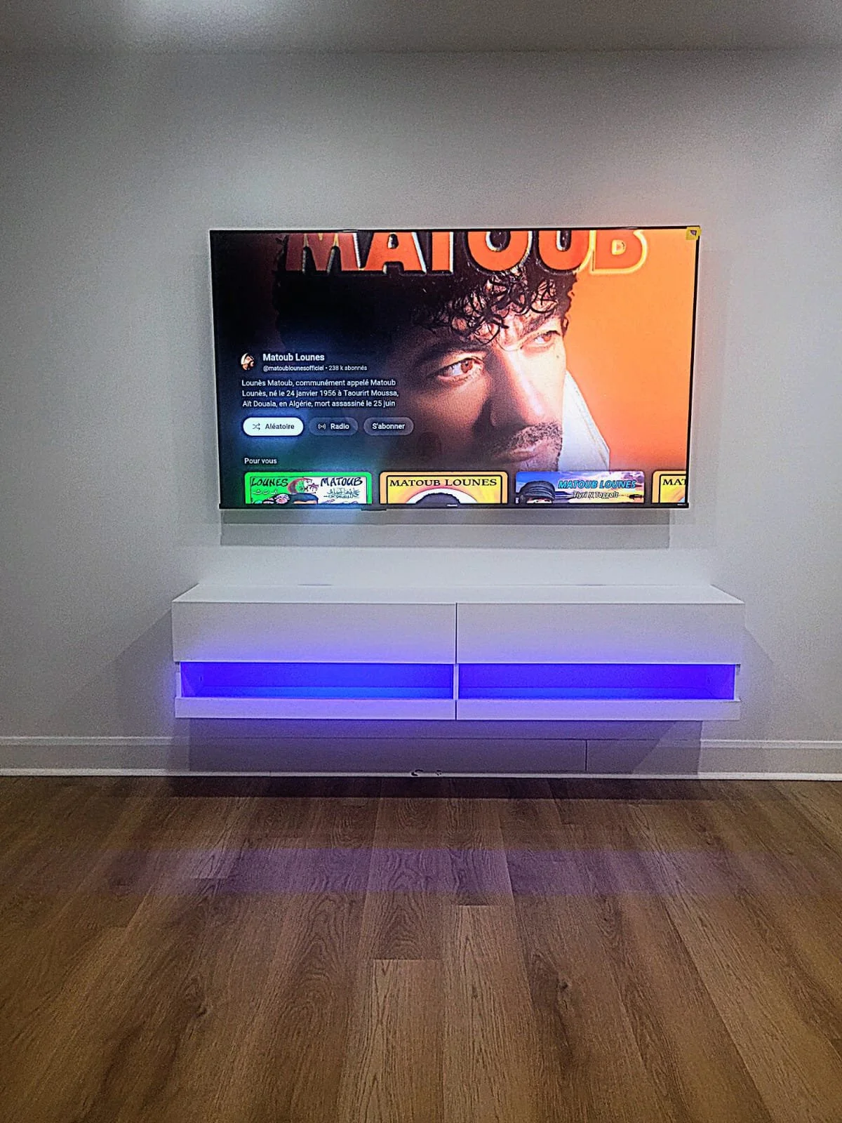 Télévision affichant une image d'un homme avec des cheveux bouclés, des textes en arabe, et plusieurs onglets de streaming ou de recommandations en bas, fixée au mur blanc avec un meuble blanc sous la télévision illuminé de lumière bleue, posé sur un