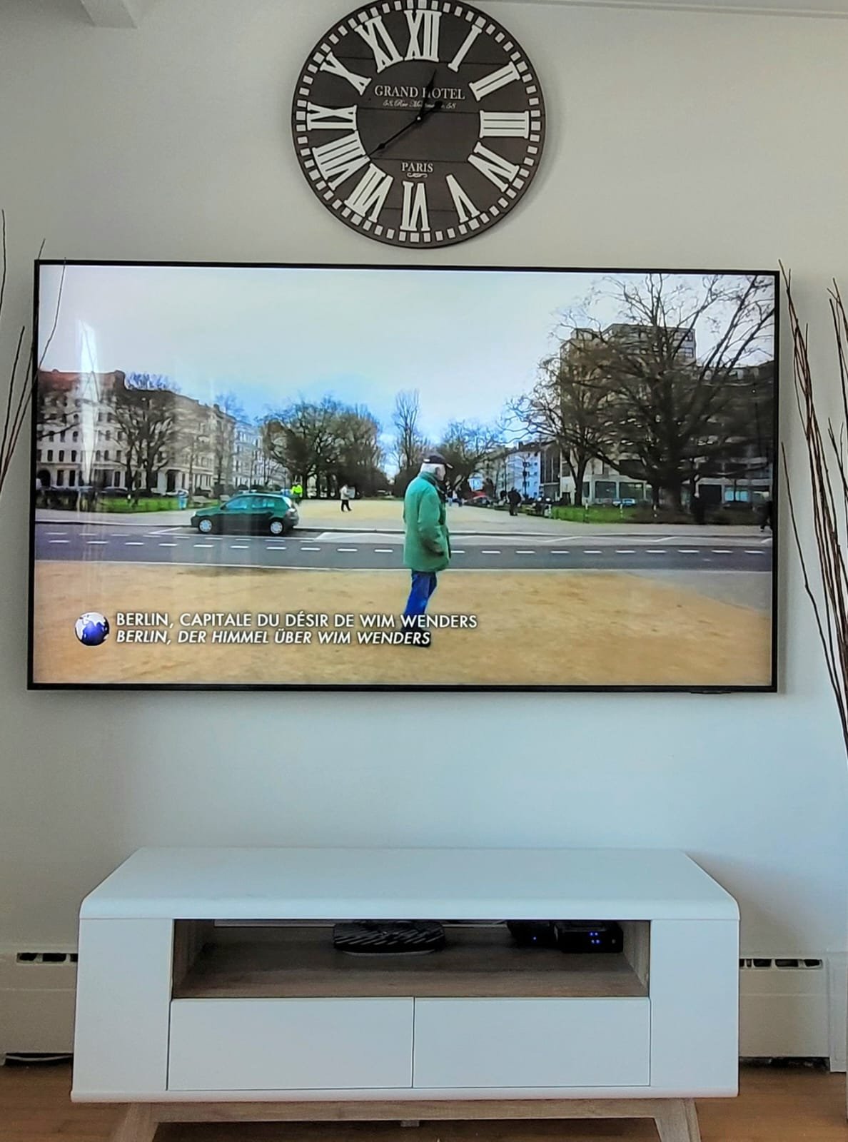 Une télévision affiche une scène en plein air à Berlin avec des personnes et des bâtiments, sous un ciel clair. Au sommet, une grande horloge murale de style vintage indique minuit.