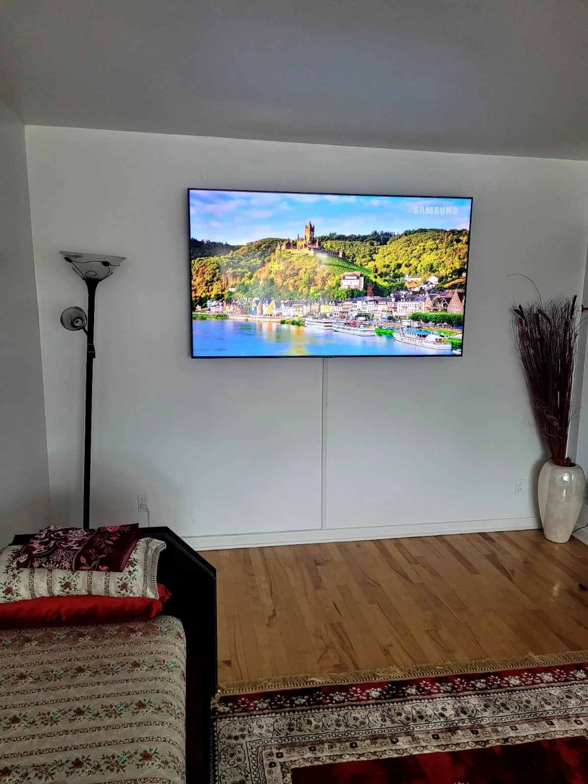 Une télévision à écran plat affichant une vue d'une ville au bord d'une rivière avec un château en haut d'une colline, dans une pièce avec un parquet en bois, un canapé avec une couverture, une lampe et un grand vase avec des branches.