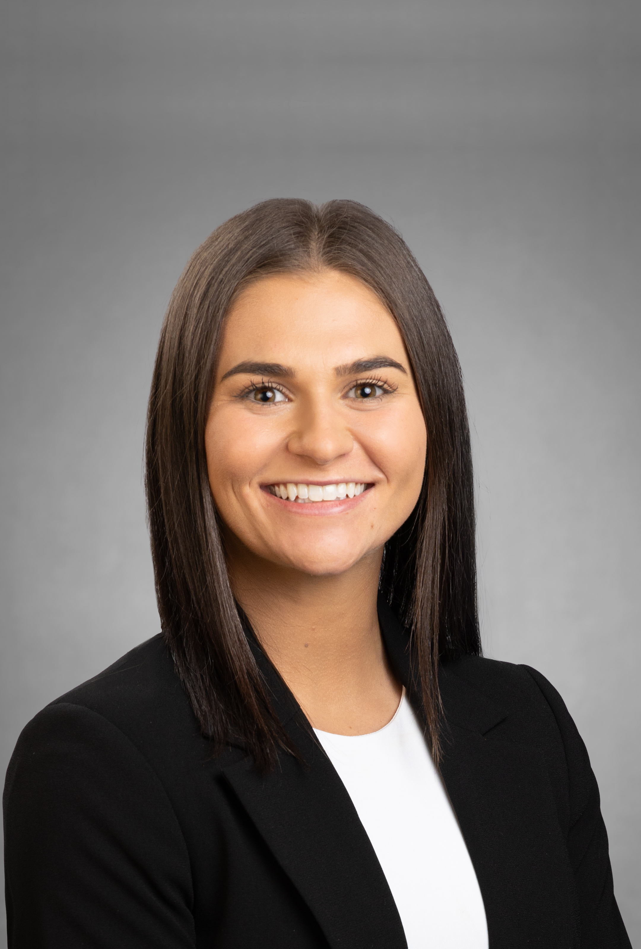 A woman with dark brown hair wearing a black blazer and white top, smiling against a gray background.