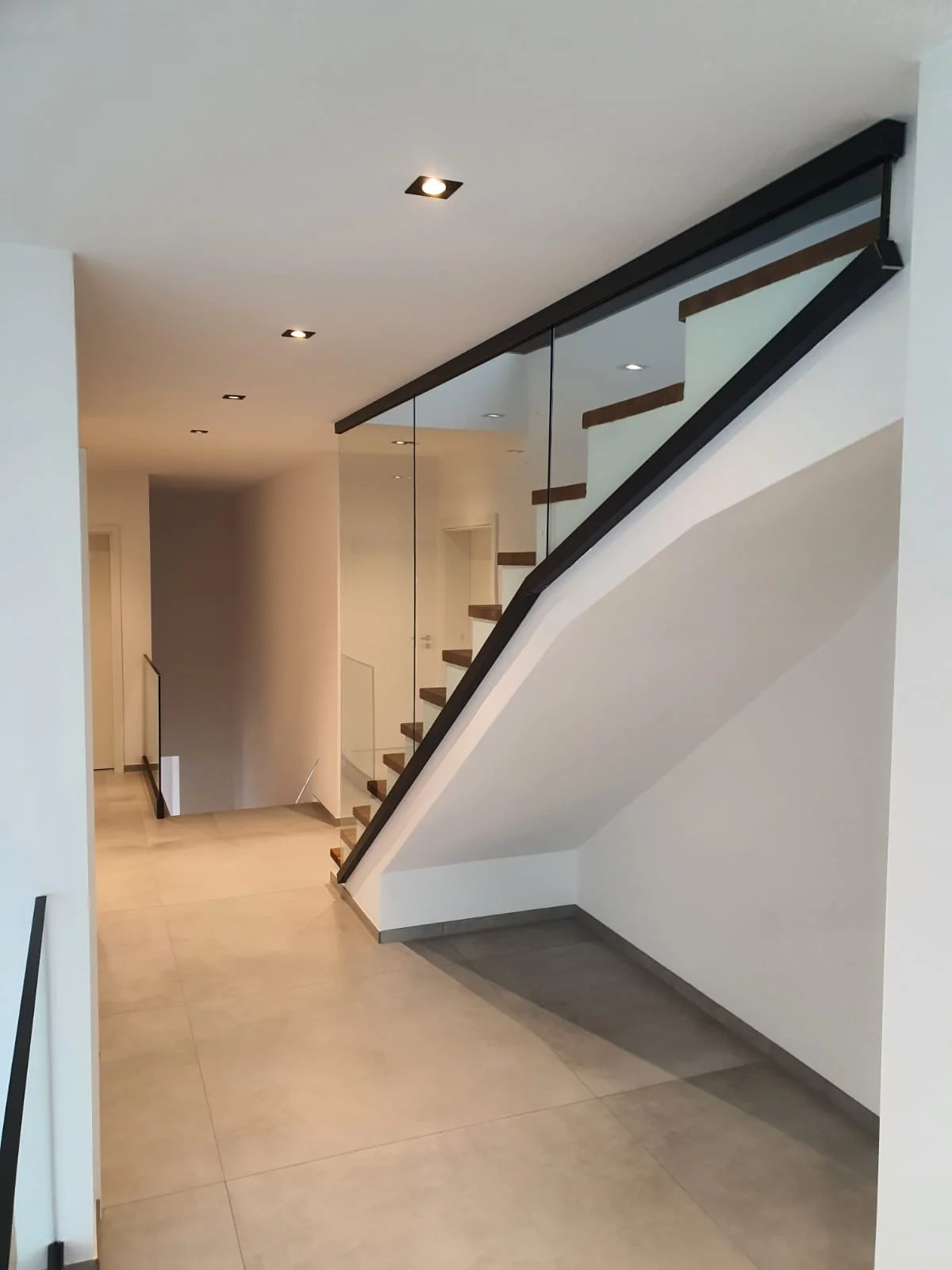 Innenaufnahme eines modernen Hauses mit Treppe, die eine Glasbalustrade hat, Laminatboden und weiße Wände, Deckenstrahler, helles Gesamtbild.