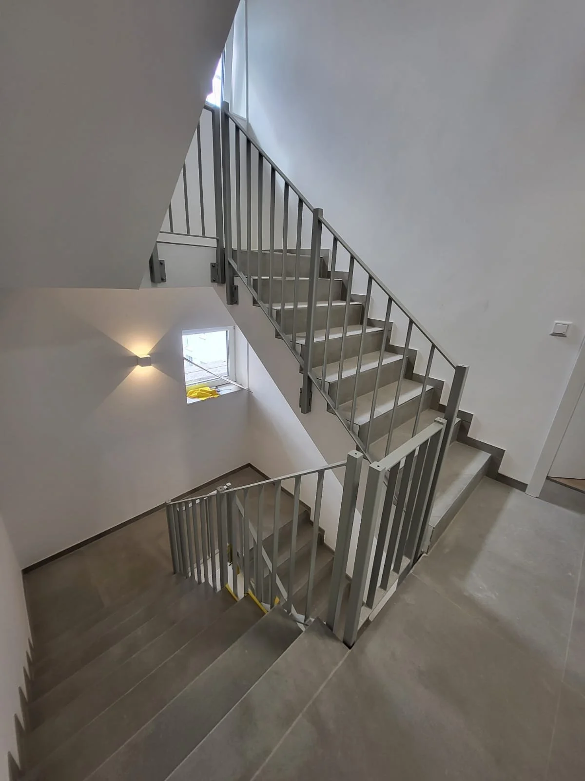 Innenansicht eines modernen Treppenhauses mit grauen Treppenstufen, einem Handlauf aus Metall und weißen Wänden, eine kleine Fensteröffnung ist sichtbar.