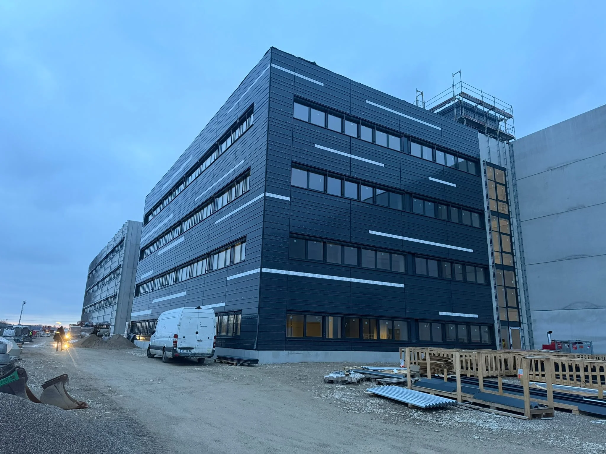 Mehrstöckiges modernes Gebäude unter Bau, mit Baugerüsten, Baustelle mit Fahrzeugen und Baugerüsten im Vordergrund, bei Sonnenuntergang.