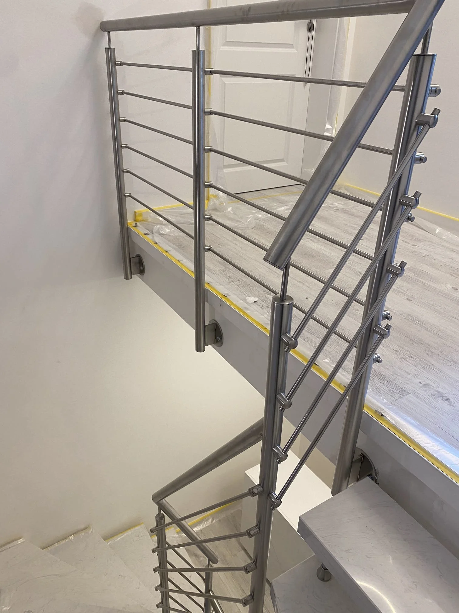Innenansicht eines neuinstallierten Edelstahlgeländers an einer Treppe in einem modern eingerichteten Raum mit weißer Wand und weißen Tür. Der Boden ist hell und die Treppe hat helle Stufen.