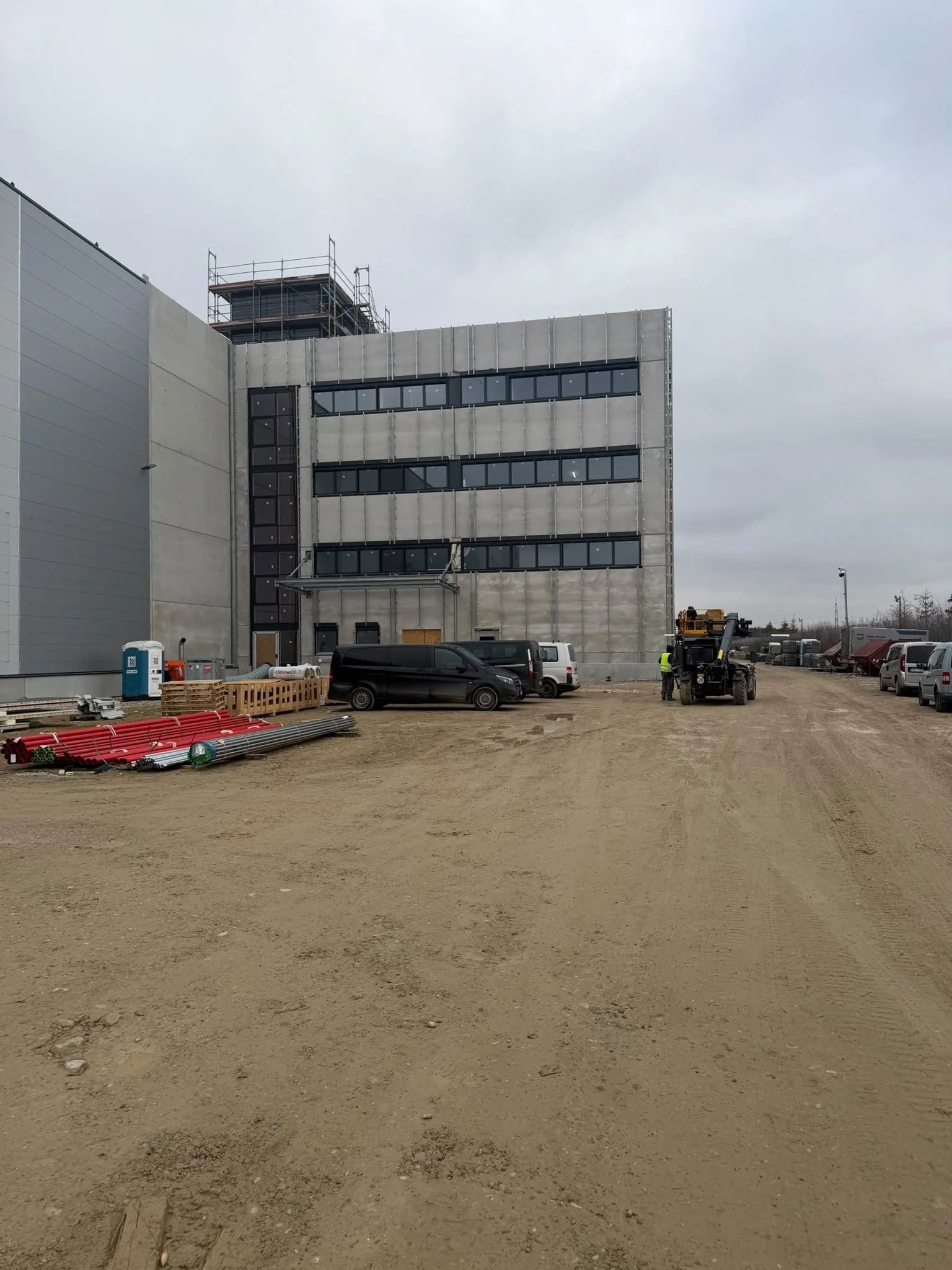 Baustelle mit einem modernen Gebäude im Bau, mit mehreren Stockwerken und Fenstern, Baustellenfahrzeugen und Baumaterialien im Vordergrund, unter einem bewölkten Himmel.