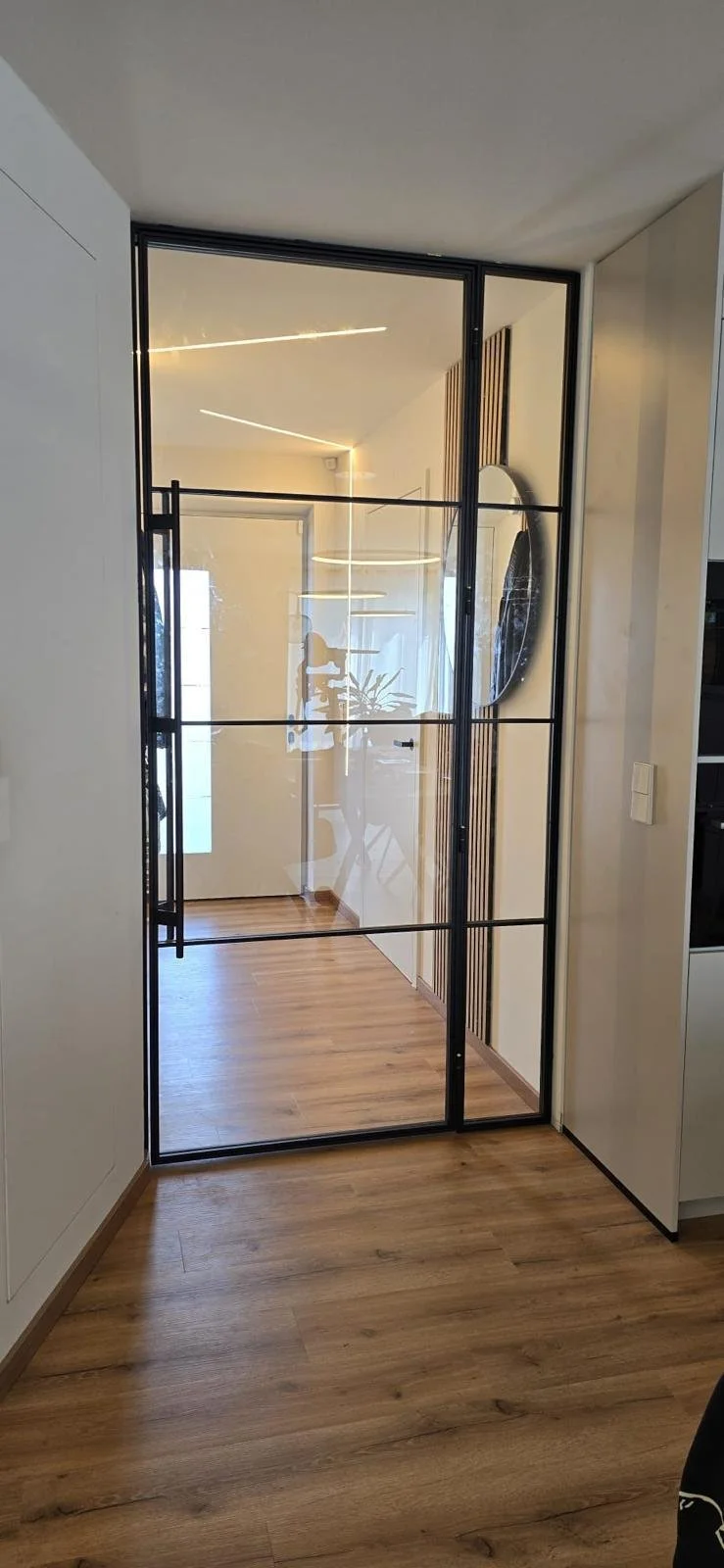 Innenansicht eines modernen Raumes mit Badezimmerausschnitt, schwarzem Metallrahmen, Holzfußboden und Spiegel an der Wand.