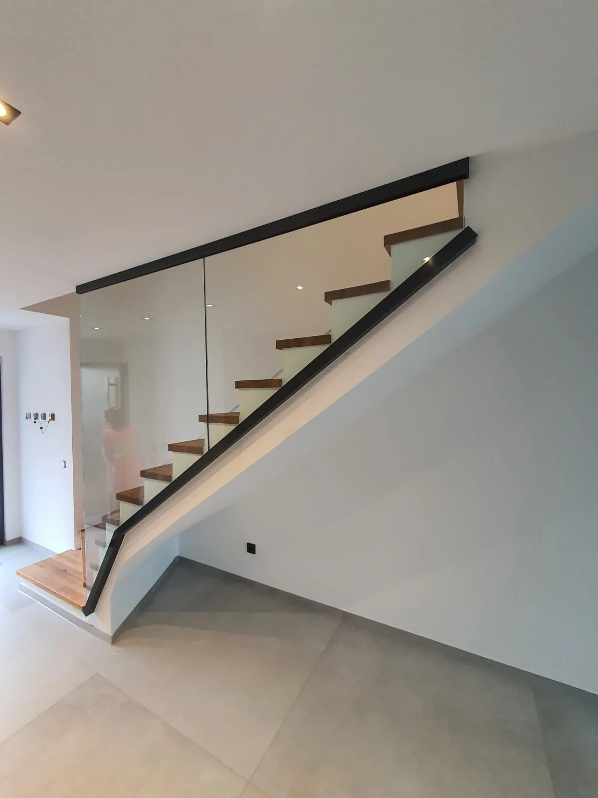 Innenraum mit moderner Treppe mit Holzstufen, Glasgeländer und schwarzem Handlauf, in einem hellen Raum mit weißen Wänden und Decke.