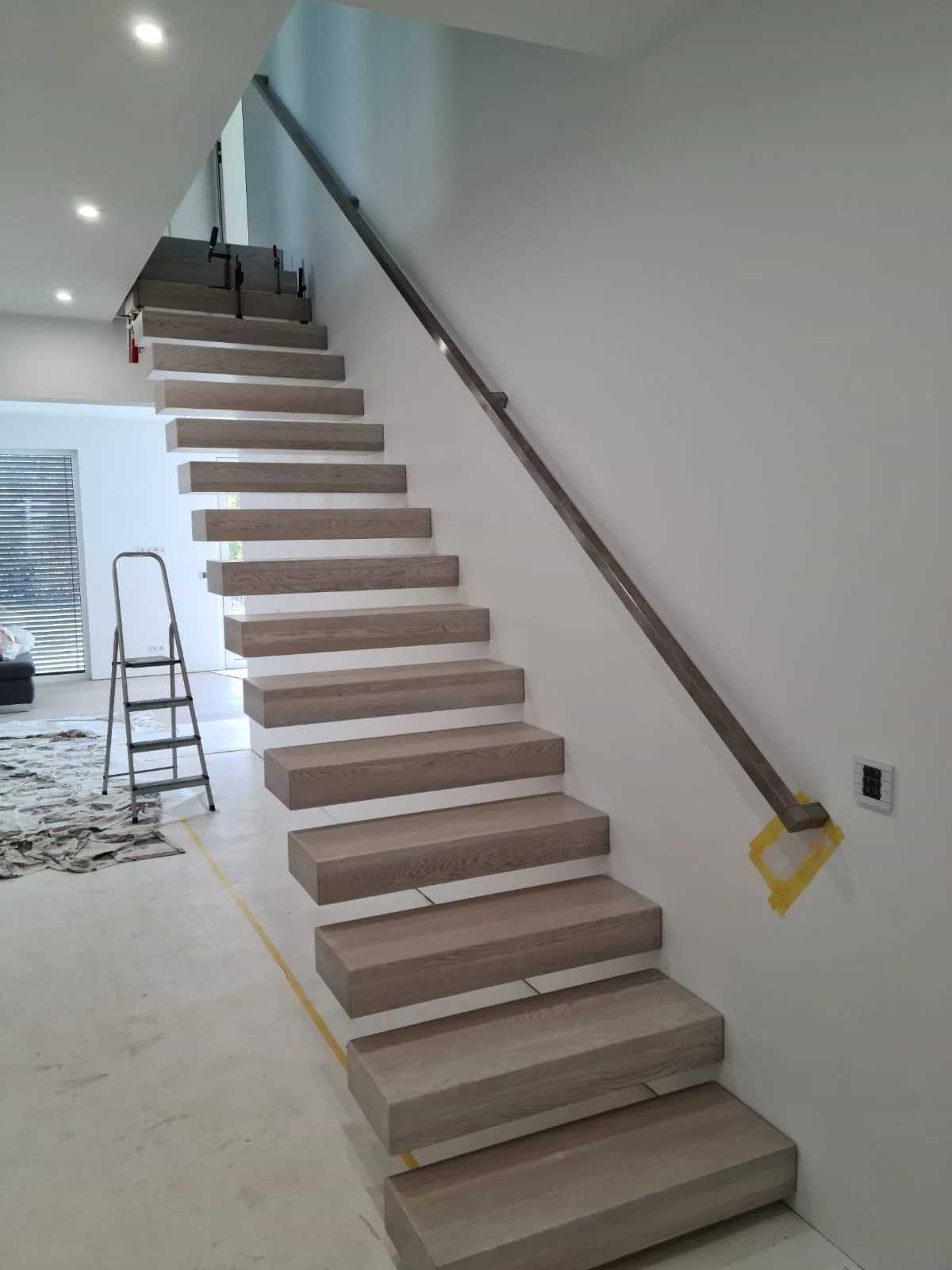 Unvollendete Innentreppe mit Holzböden, Geländer und Installationen, im Hintergrund ein Raum mit einem Sofa und einem Fenster mit Jalousien.