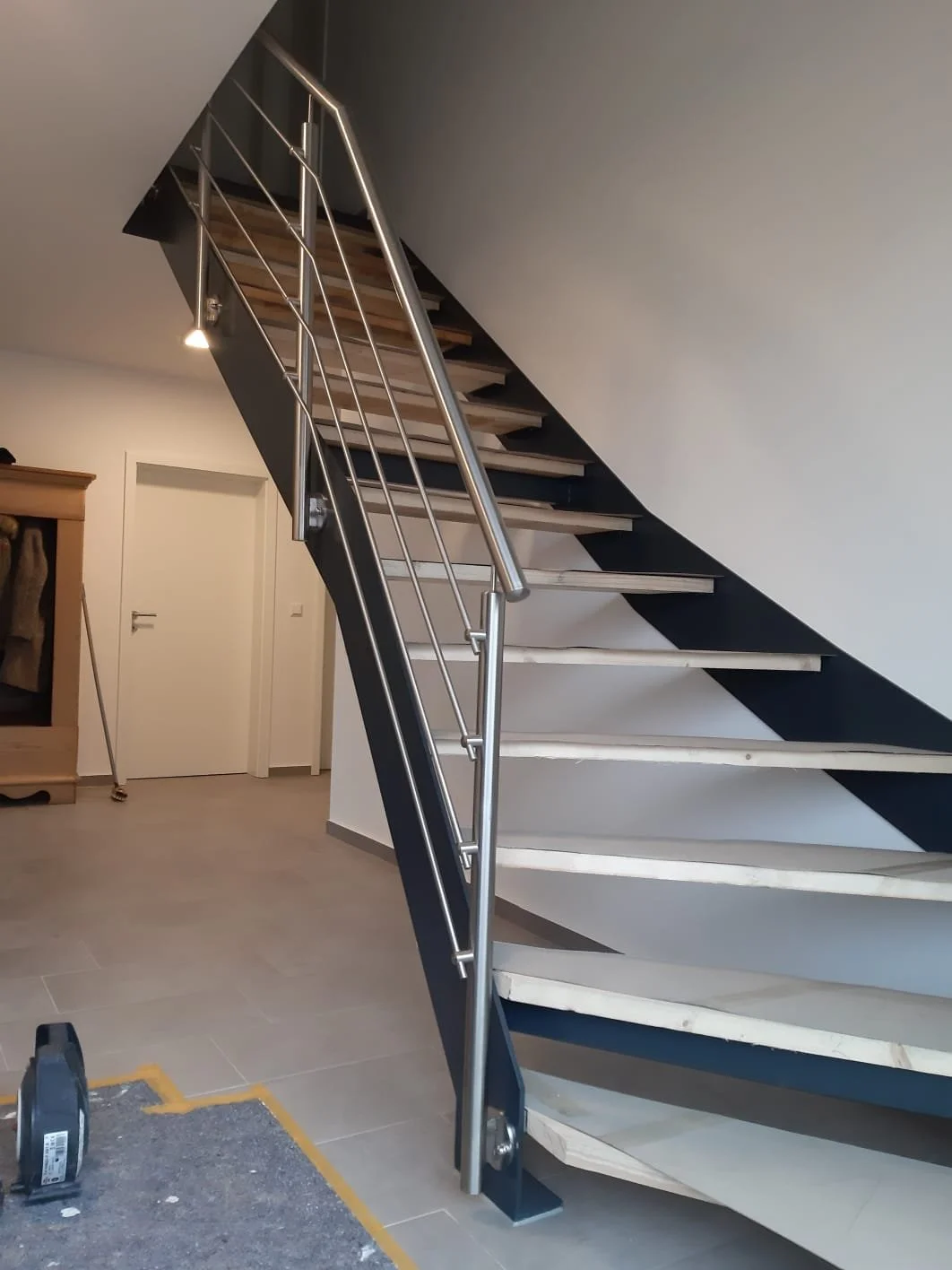 Innenansicht einer halb fertigen Treppe mit Holzstufen und Edelstahlhandlauf in einem Zimmer mit weißen Wänden.