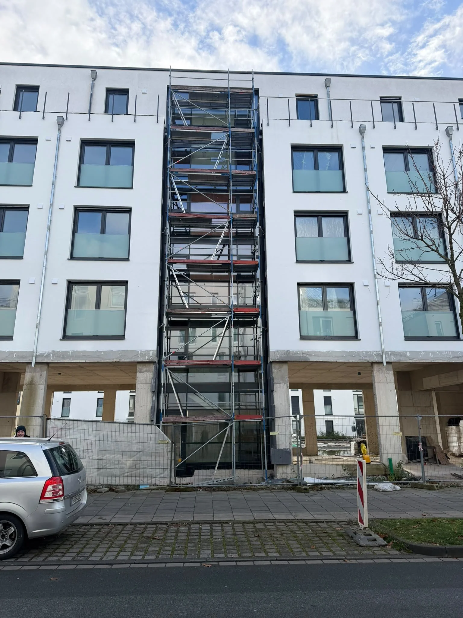 Mehrstöckiges Baugerüst vor einem weißen modernen Gebäudekomplex unter blauem Himmel mit Wolken. Im Vordergrund sind ein parkendes Auto und ein Gehweg mit Ziegelsteinen sichtbar.