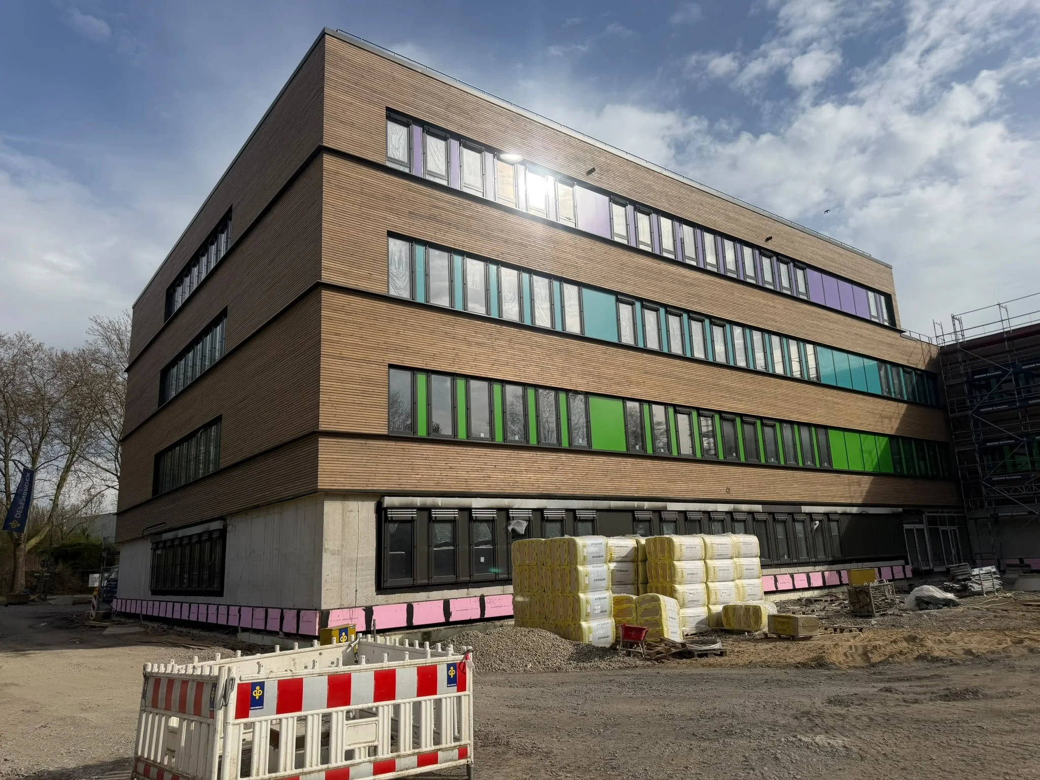 Mehrstöckiges Gebäude unter Bau mit Holzfassade, große Fenster, Baustellenmaterialien und Absperrung im Vordergrund.