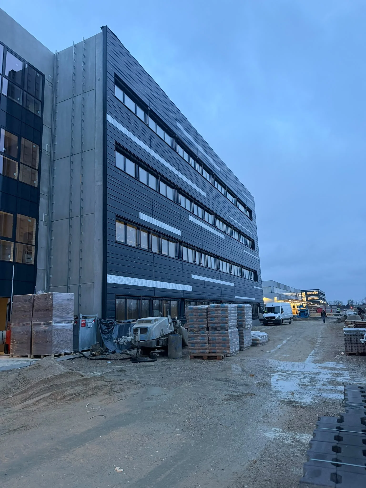 Baustelle vor einem modernen Bürogebäude mit dunkler Fassade, Baumaterialien und Maschinen im Vordergrund, Menschen und weitere Gebäude im Hintergrund.