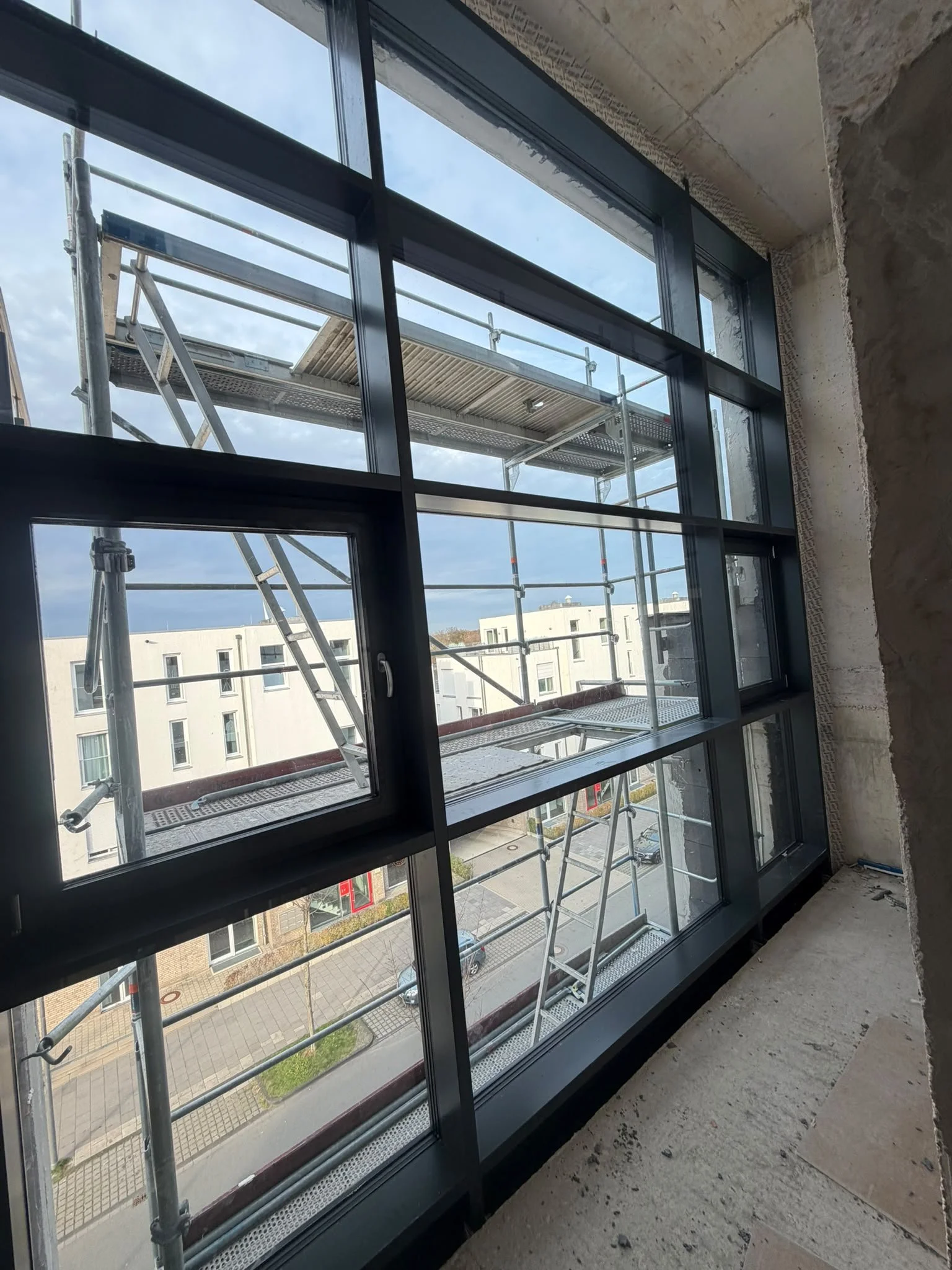 Innenansicht eines Gebäudes mit Blick auf ein großes Fenster und Gerüst außen, Aussicht auf eine Wohnanlage mit weißen Mehrfamilienhäusern und Straßen. Der Raum ist unvollendet mit Betonboden und unverputzter Wand.
