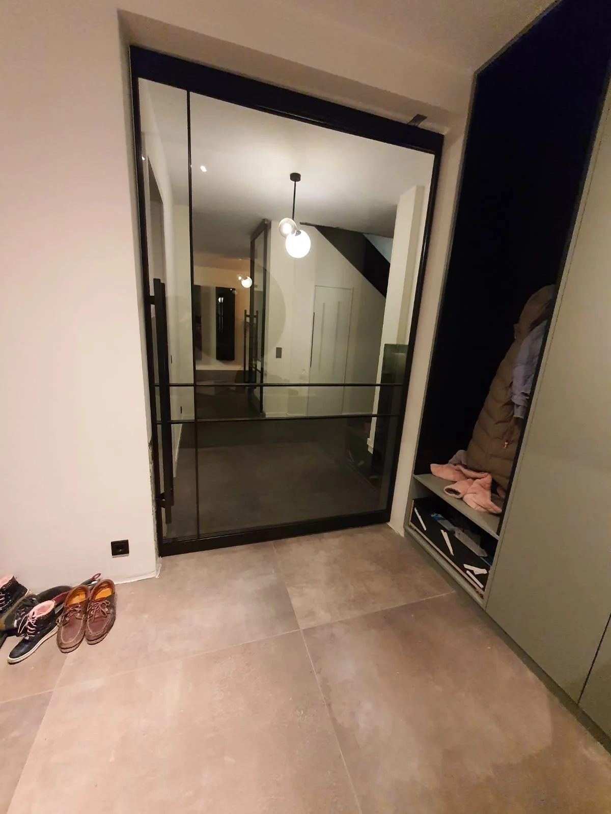 Eingangsbereich mit Fliesenboden, Schuhe auf dem Boden, eine Glas-Schiebetür, die in einen Flur führt, und eine Garderobe mit Mänteln und Schuhen