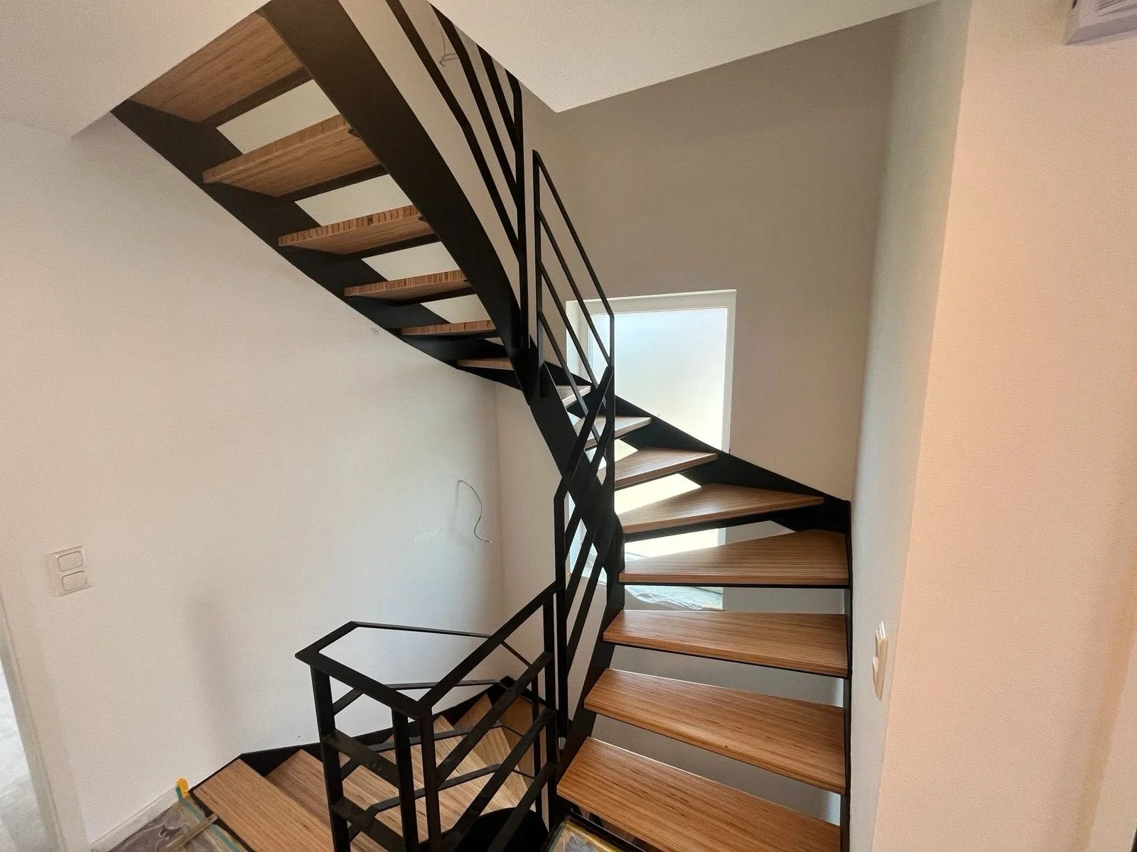Innenansicht einer modernen spiralförmigen Holztreppe mit schwarzen Metallgeländern, die in einem hellen Raum mit weißer Wand und einem kleinen Fenster führt.
