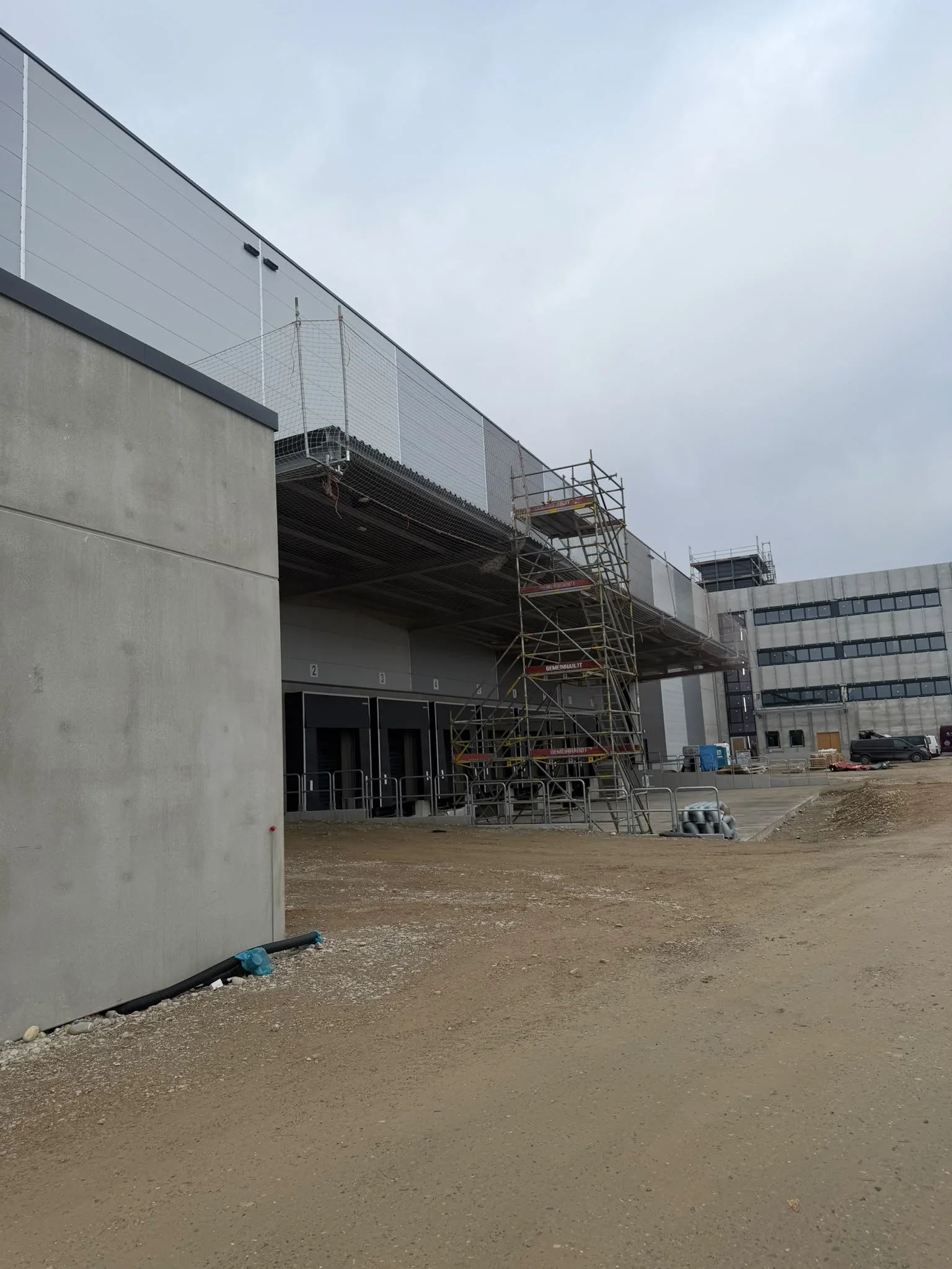 Baugerüst an einer modernen Gebäudefront unter grauem Himmel, Baustelle mit unbefestigtem Boden und Rohren im Vordergrund.