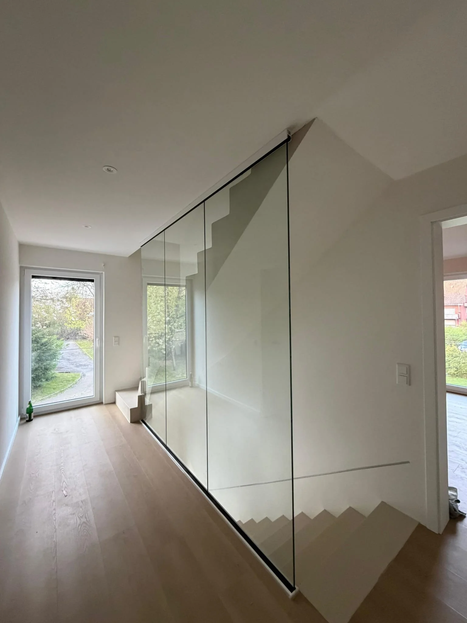 Innenaufnahme eines modernen Hauses mit Treppe, große Fenster, glatter Boden und eine Glastür, im Hintergrund nach draußen blickend auf Bäume und Natur.