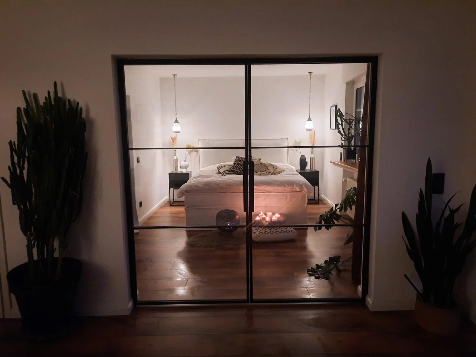 Ein Schlafzimmer gesehen durch eine Glastür, beleuchtet mit warmem Licht, mit einem Bett, Bettrahmen, Nachttischen, Lampen, Deckenventilator, Pflanzen und Kerzen.