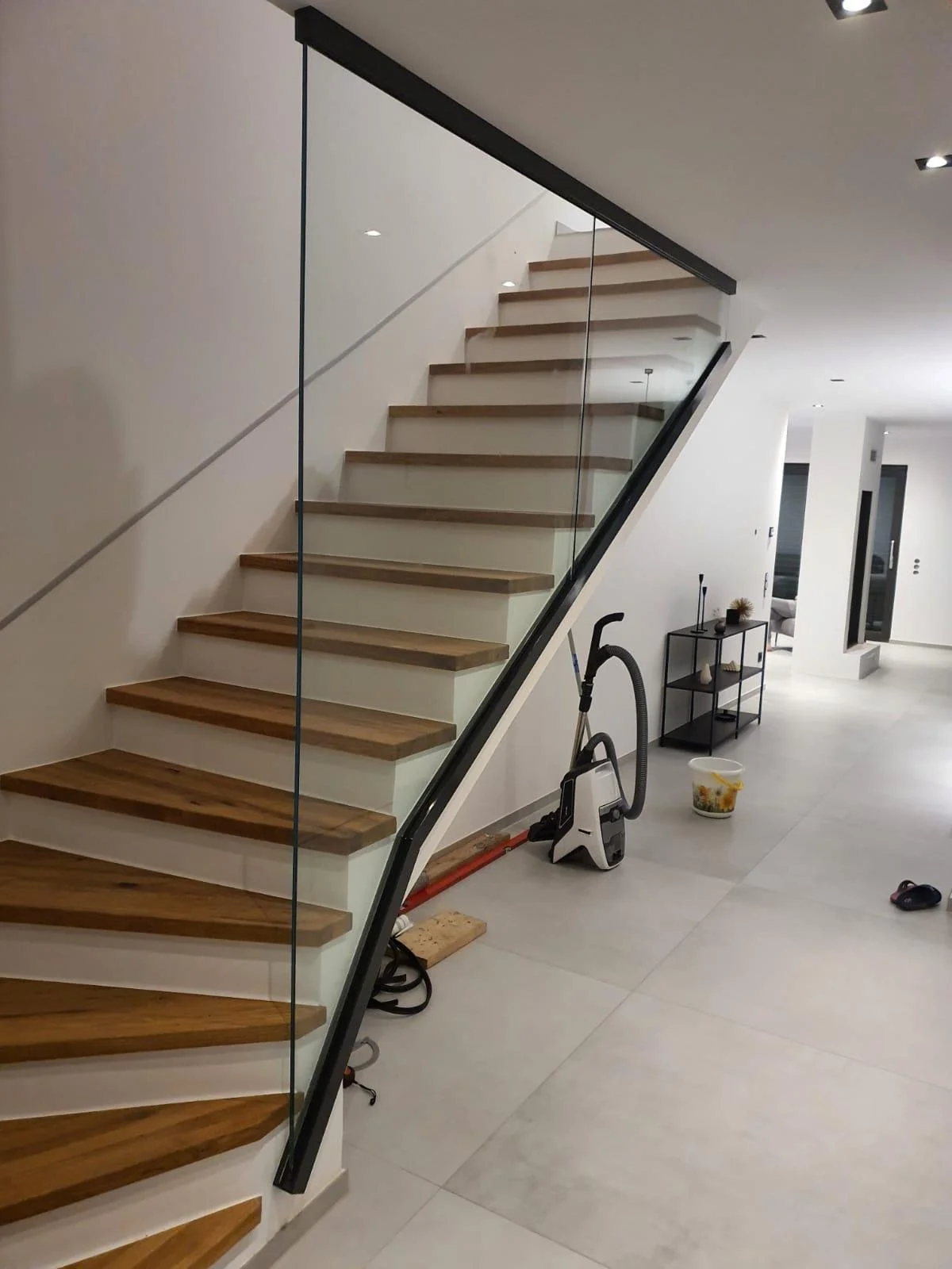Innenansicht eines modernen Hauses mit Treppe, Bodenstaubsauger, einer kleinen schwarzen Konsole mit Dekoration, weißen Wänden und großen Fenstern.
