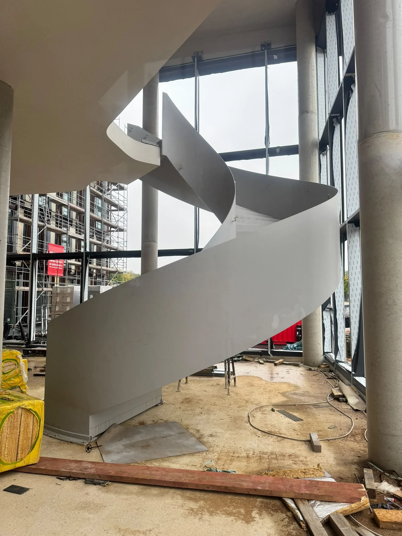 Innenansicht eines Gebäudes unter Bau, mit einer spiralförmigen weißen Treppe, Bauarbeiten und Baumaterialien im Vordergrund, offene Gitterfenster und Baugerüst im Hintergrund.