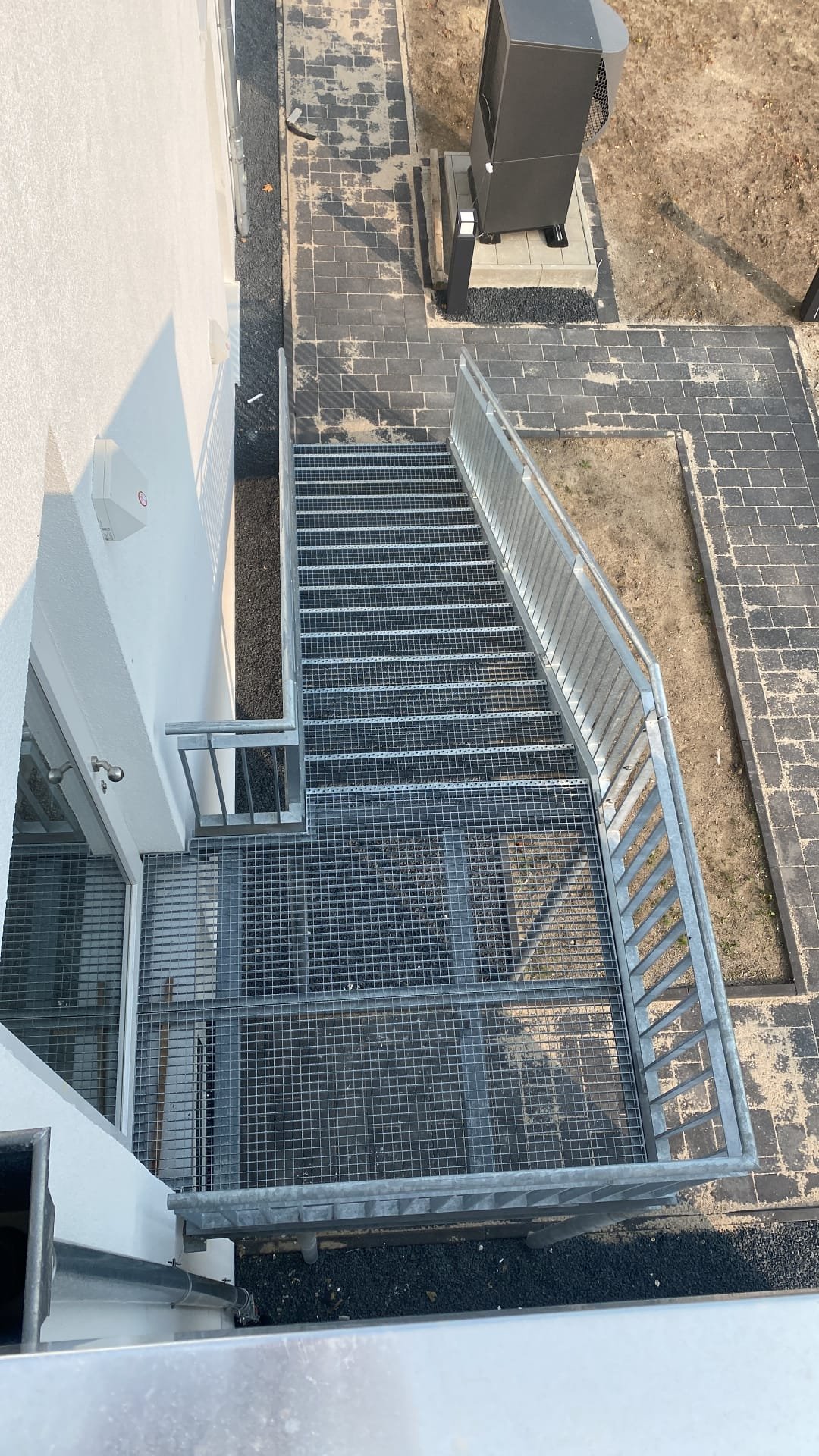 Blick auf eine Metalltreppe vor einer weißen Wand, mit einem elektrischen Kabel und einem Boden aus schwarzen und grauen Fliesen, daneben eine unbefestigte Erdfläche.