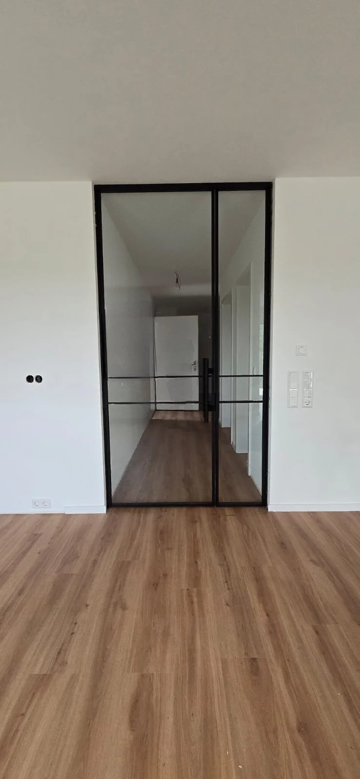 Innenraum eines modernen Hauses mit Holzfußboden und Glastür, Zugang zu einem Flur im Hintergrund.