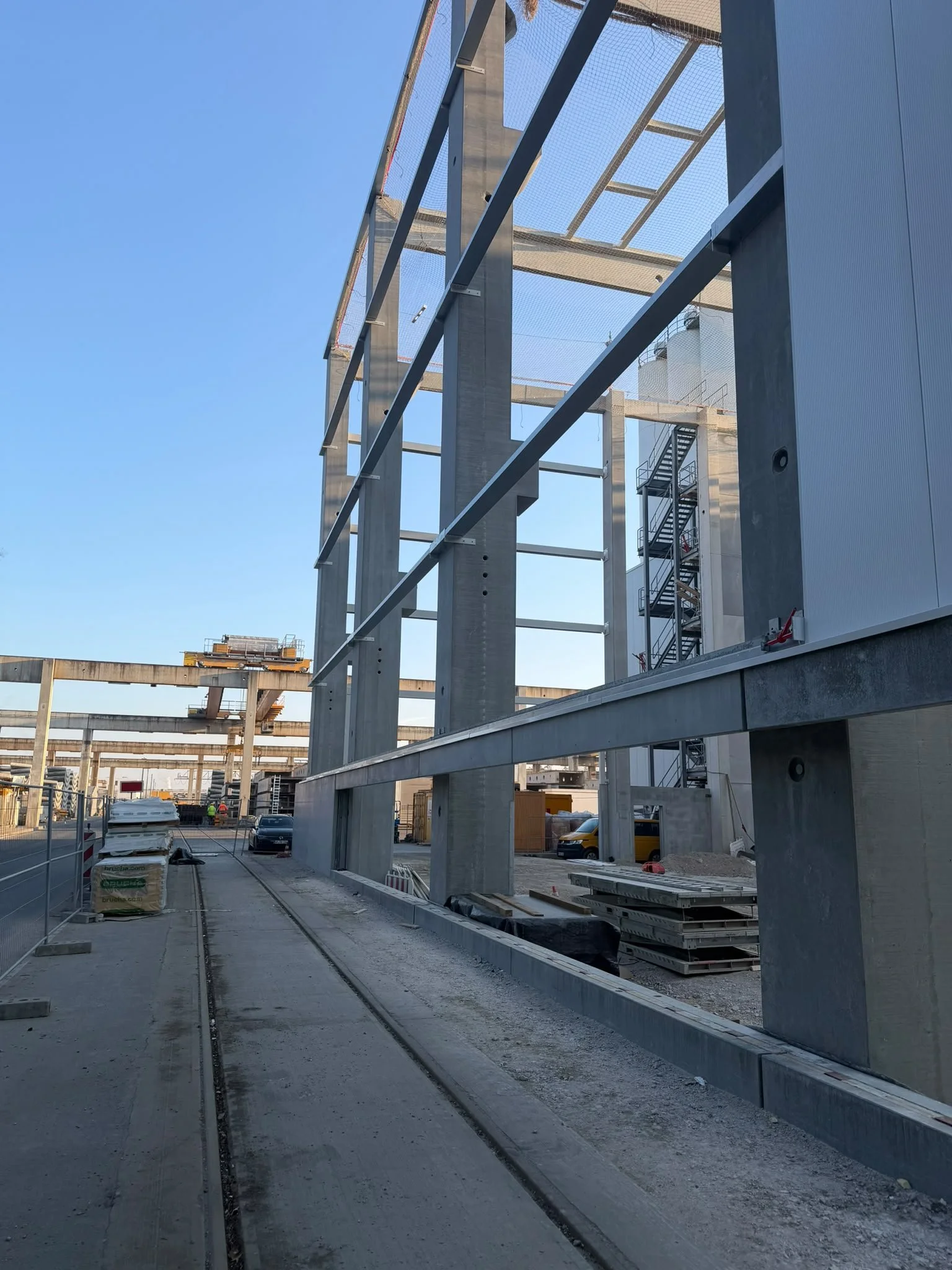 Baustelle mit Stahlkonstruktion und Baugerüsten, Bauarbeiter bei der Arbeit, Versorgungsfahrzeuge im Hintergrund, blauer Himmel.