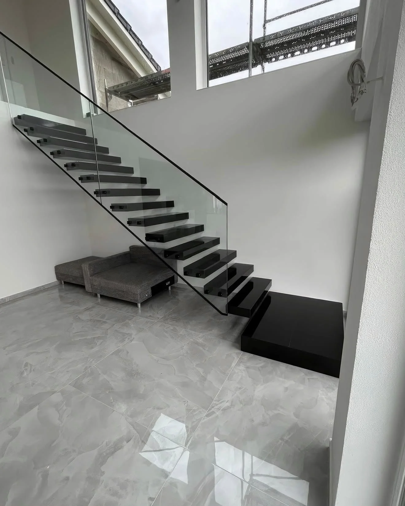 Moderne schwarze Treppe mit Glasgeländer in einem hellen Raum, darunter ein graues Sofa, große Fliesen auf dem Boden, und ein Fenster mit herabgelassener Baugerüst im Hintergrund.