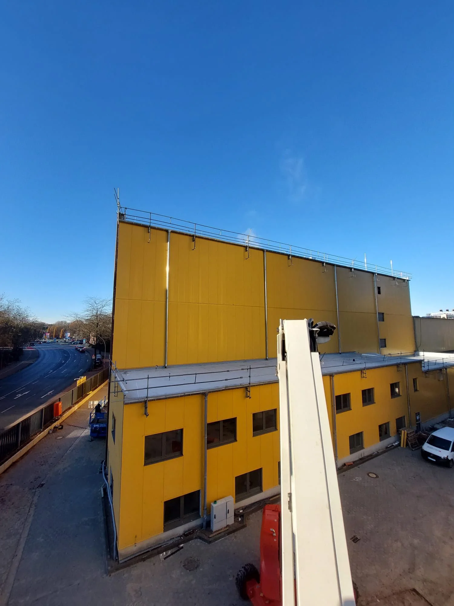 Ein gelb gestrichenes Gebäude mit mehreren Etagen und großen Fenstern. Vor dem Gebäude befinden sich Baustellenfahrzeuge und -geräte, darunter ein Arbeitsmammut. Der Himmel ist klar und blau.