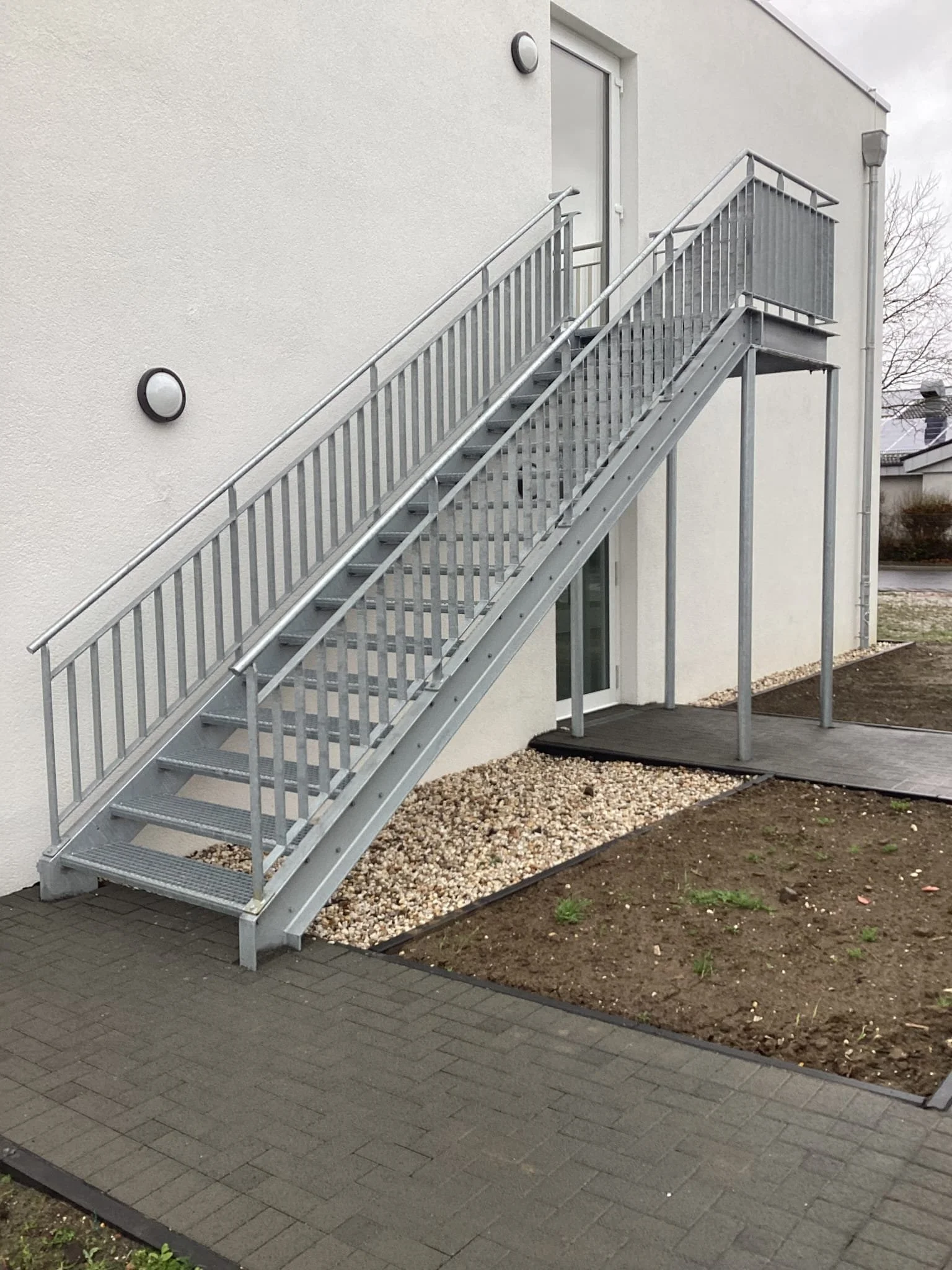 Blick auf eine Außentreppe mit Geländer neben einem modernen weißen Gebäude, darunter ein gepflasterter Weg und ein befestigter Gehweg.