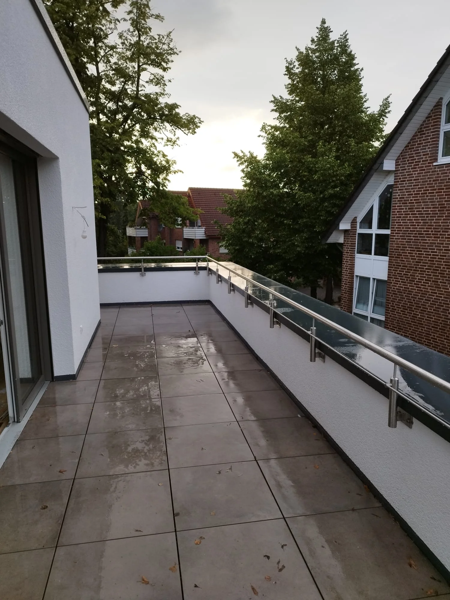 Ein leerer Balkon mit nassen Fliesen, umgeben von weißen und roten Backsteinwänden, mit Blick auf bewaldete Häuser und Bäume bei bewölktem Himmel.