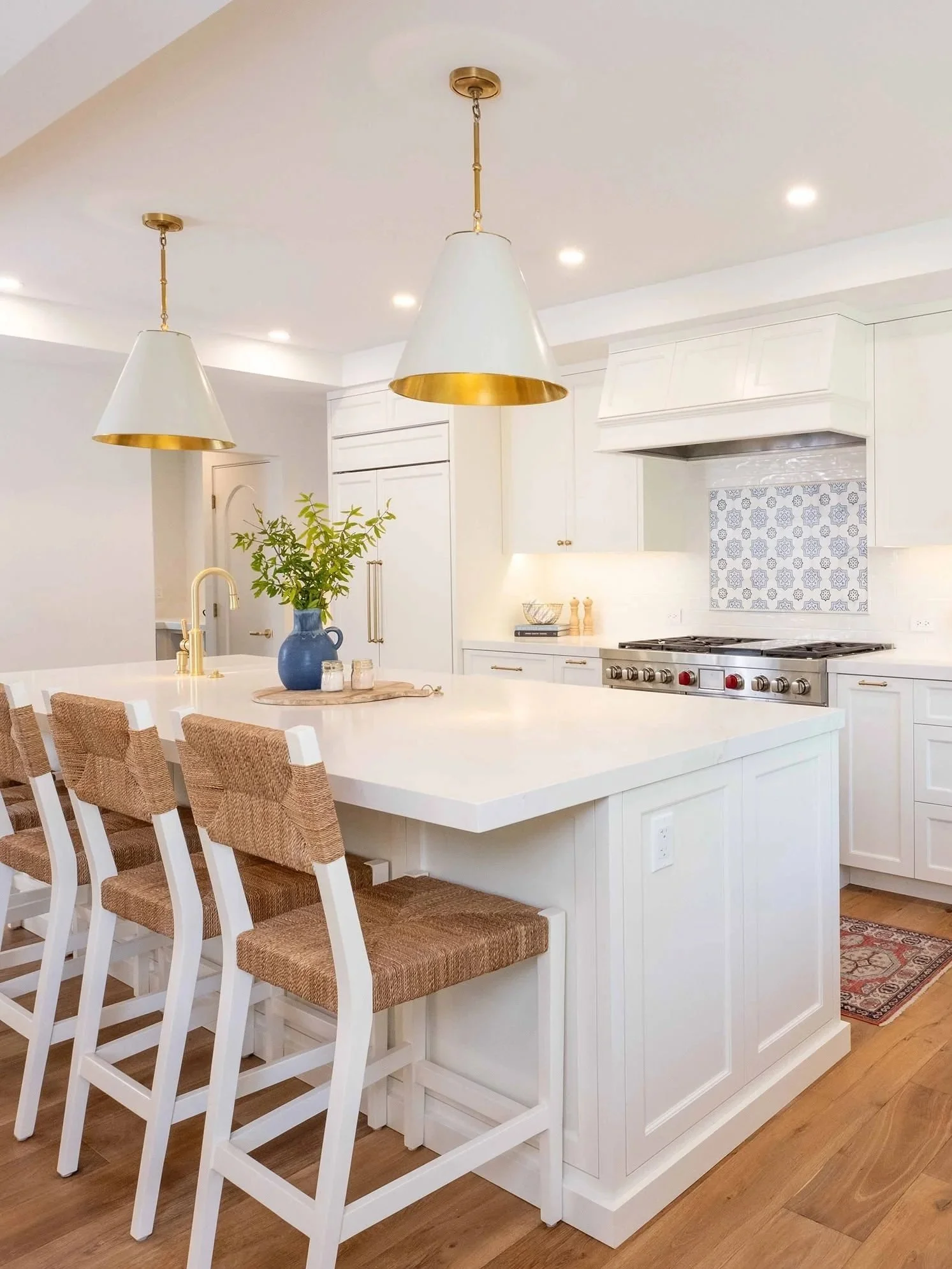Bright modern kitchen with white cabinetry, stainless steel stove, and large white island with woven-back chairs. Two gold and white pendant lights hang above the island, and a bowl with green leaves is on the counter.