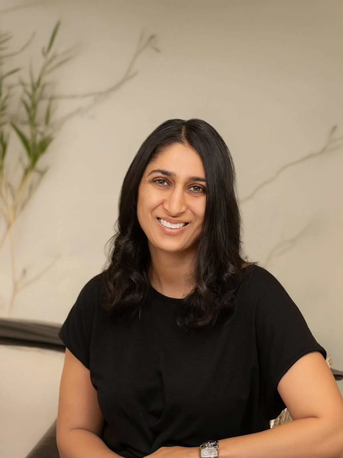 A woman with black hair and a black shirt smiling while sitting indoors with a beige wall and a tall plant in the background.