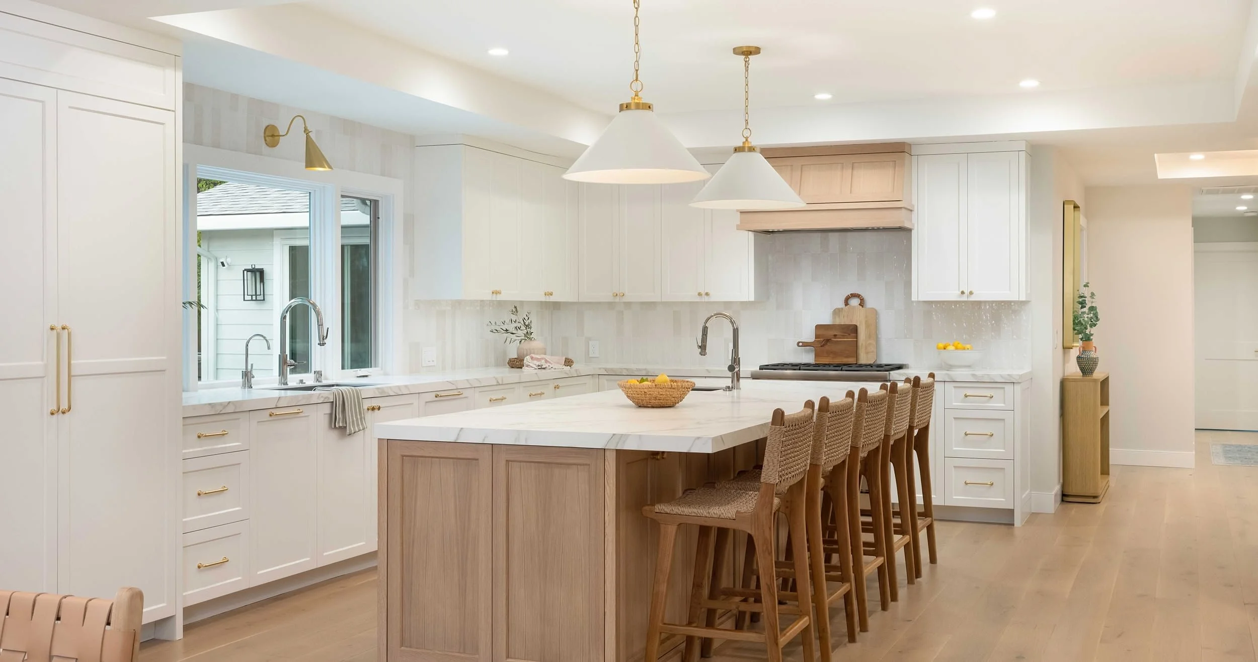 Bright, modern kitchen with white cabinetry, marble countertops, and a central island with wooden seating. Features include large windows, pendant lighting, and decorative accents like vases and bowls.